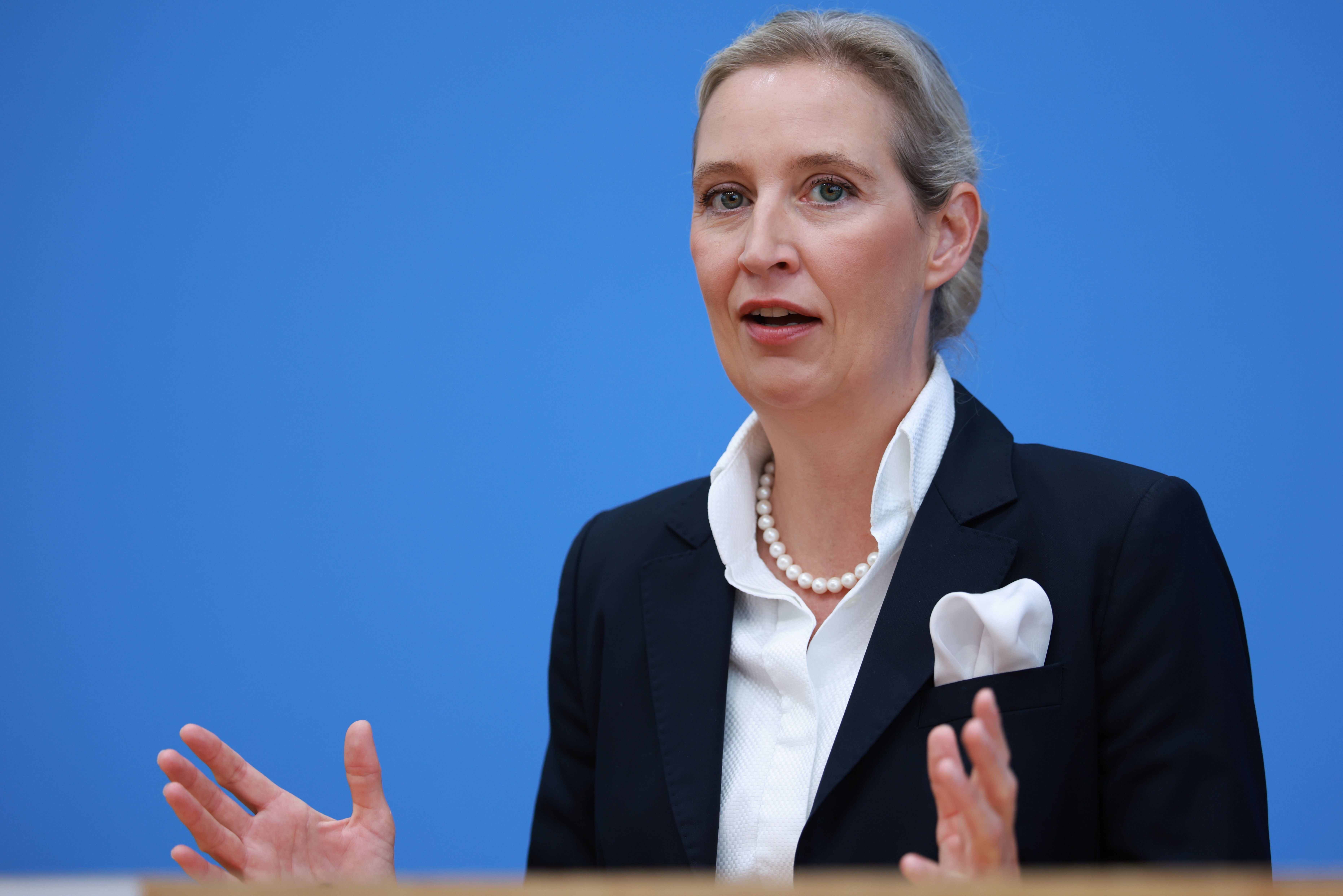 Eine Frau im Anzug spricht gestikulierend auf einer Pressekonferenz vor blauem Hintergrund.