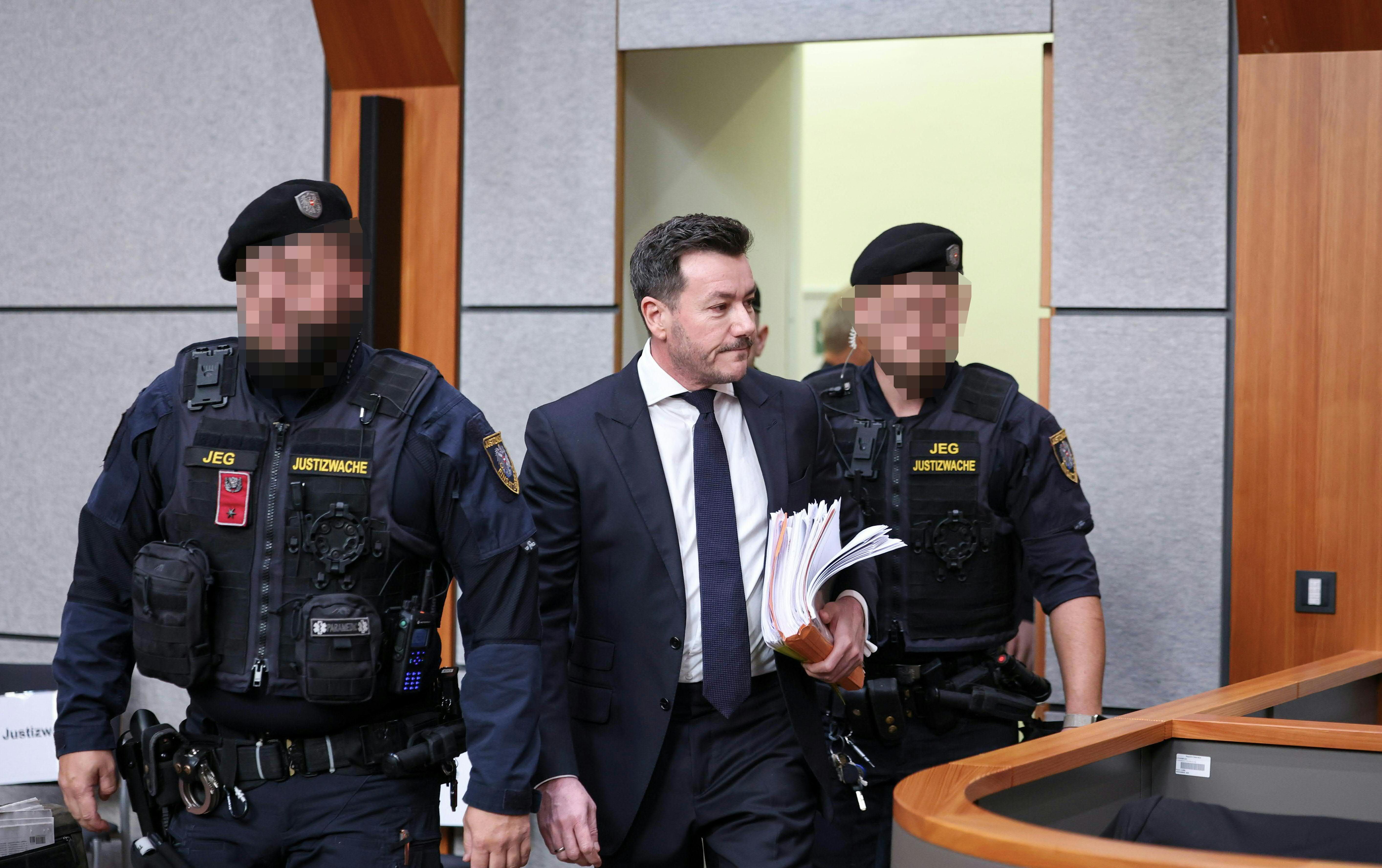Ein Mann im Anzug wird von zwei Justizwachebeamten durch einen Gerichtssaal begleitet.