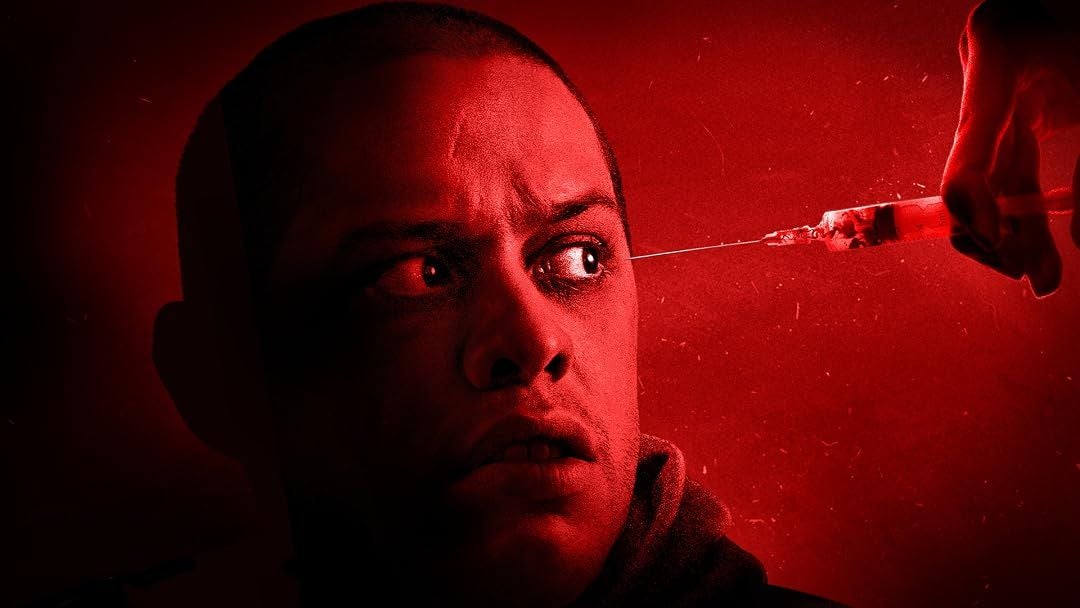 Mann blickt erschrocken auf eine Spritze, die auf sein Gesicht gerichtet ist, rotes Licht dominiert.