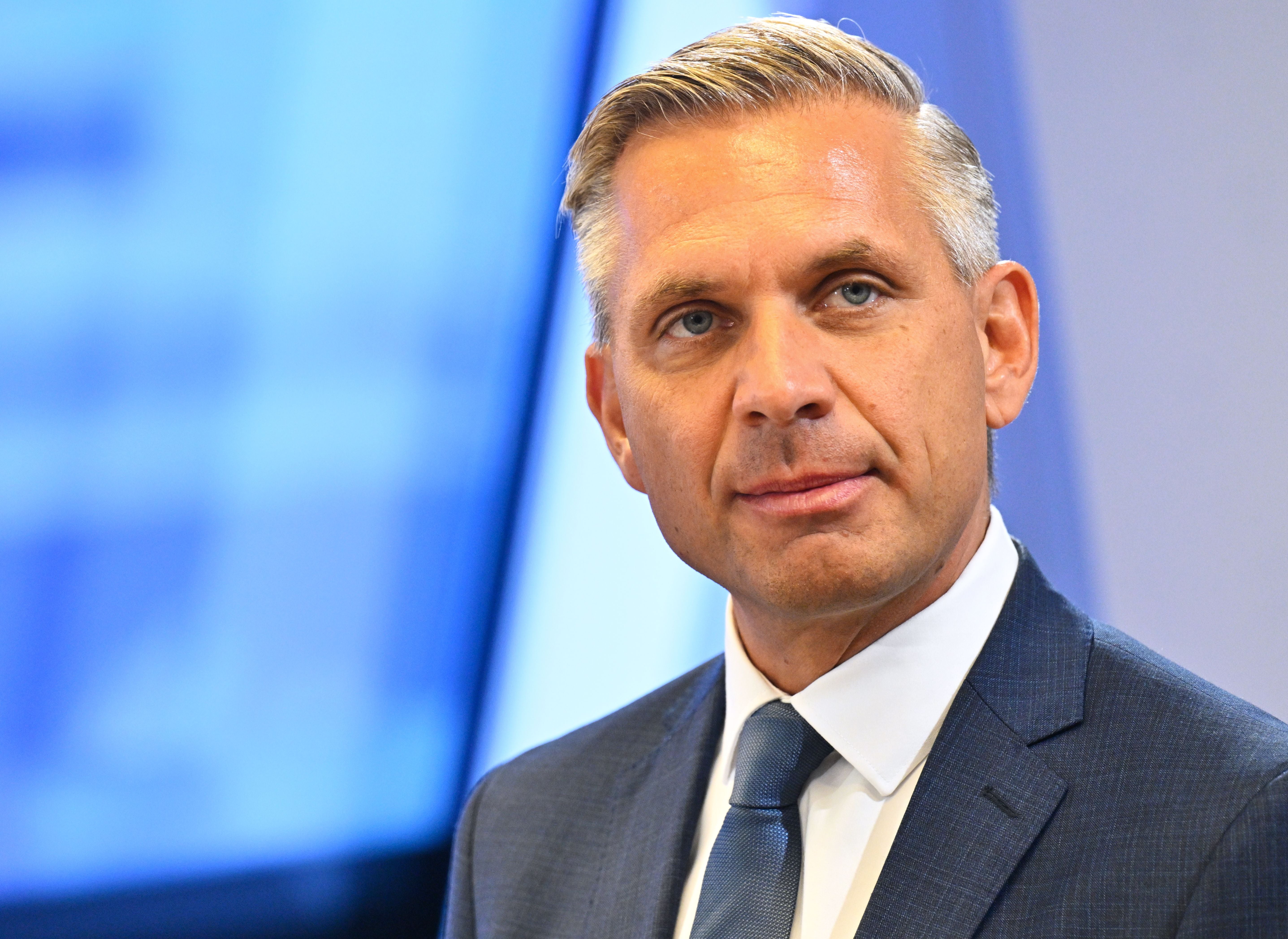 Ein Mann im Anzug mit Krawatte vor einem unscharfen blauen Hintergrund.