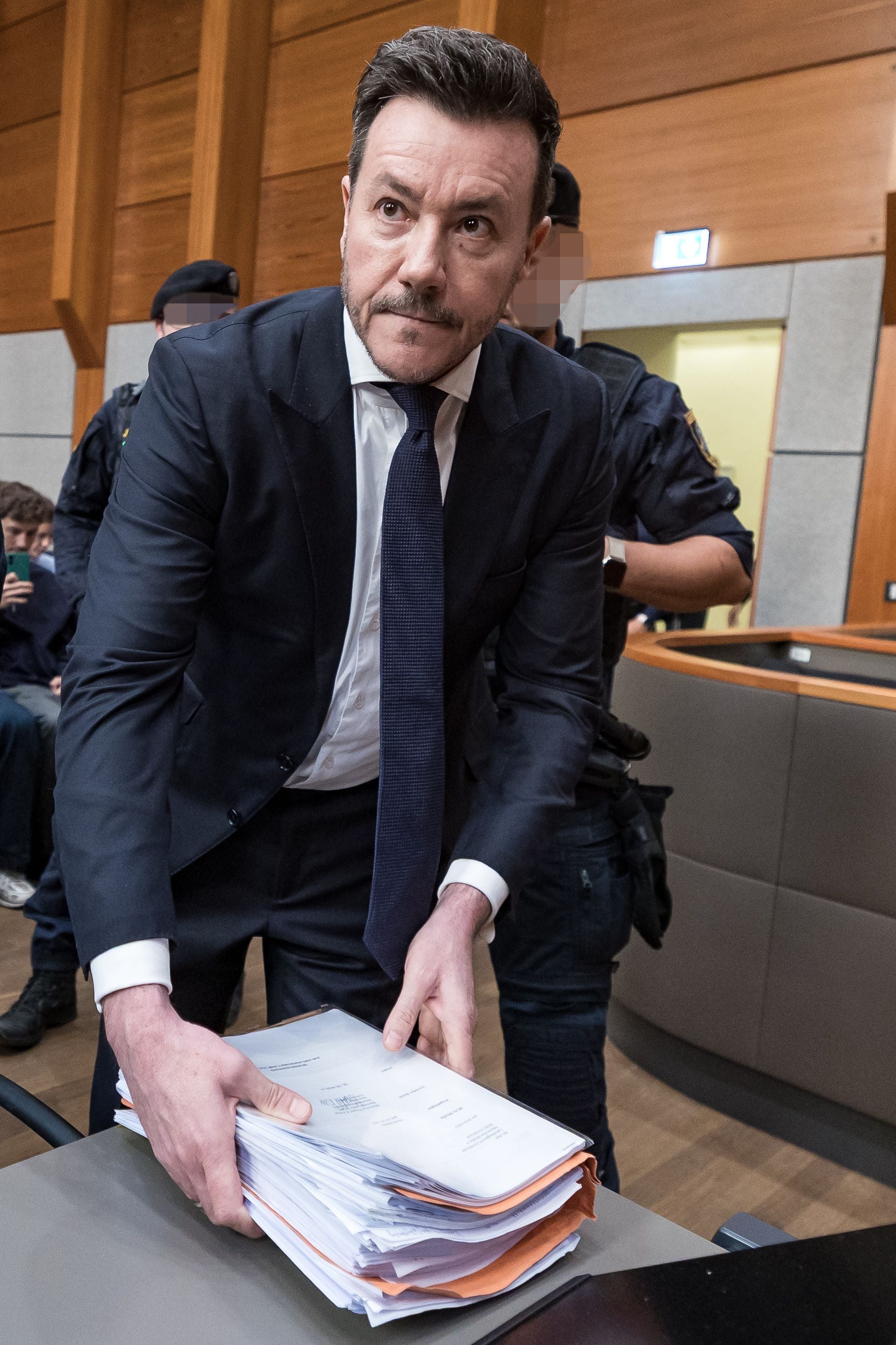 Ein Mann im Anzug legt einen Aktenstapel auf einen Tisch in einem Gerichtssaal ab.