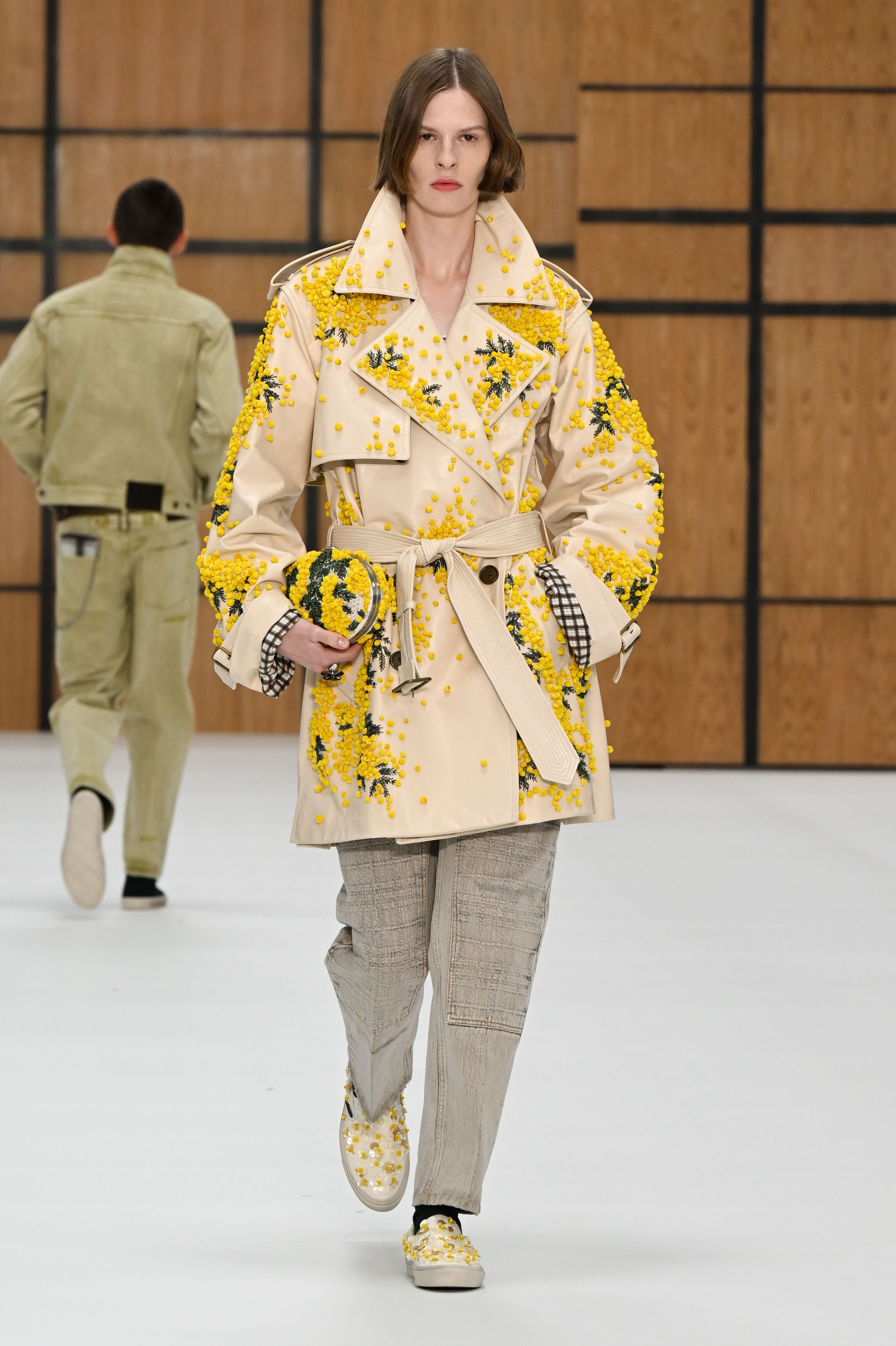Model mit beigefarbenem Mantel mit gelben Blumenapplikationen auf dem Laufsteg.