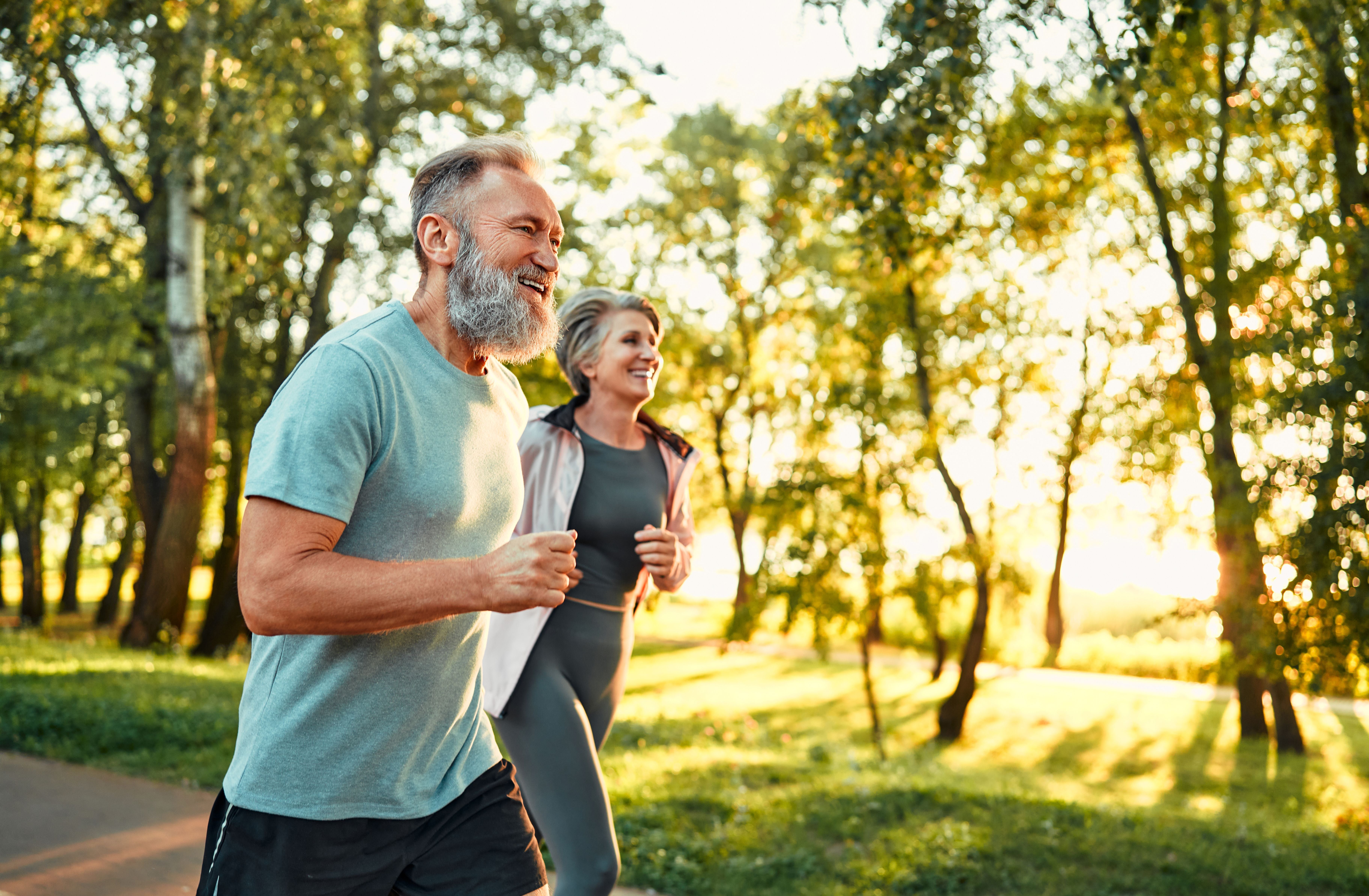 Älteres Paar joggt fröhlich im sonnigen Park, umgeben von grünem Bäumen.