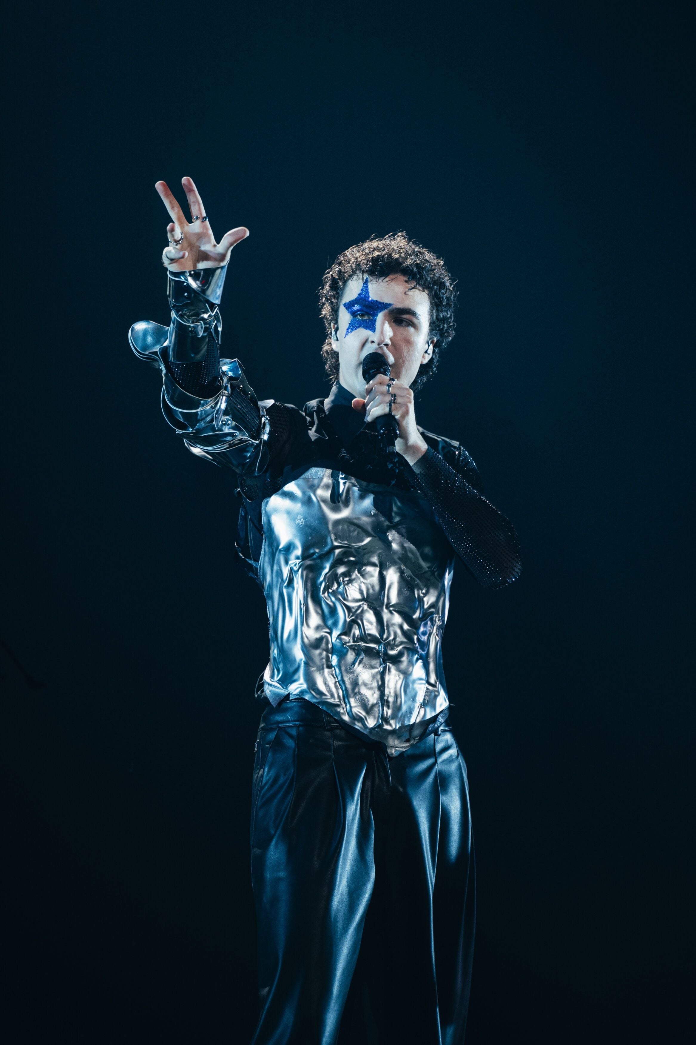 Ein Sänger mit silbernem Outfit und blauem Stern im Gesicht steht auf der Bühne und singt.