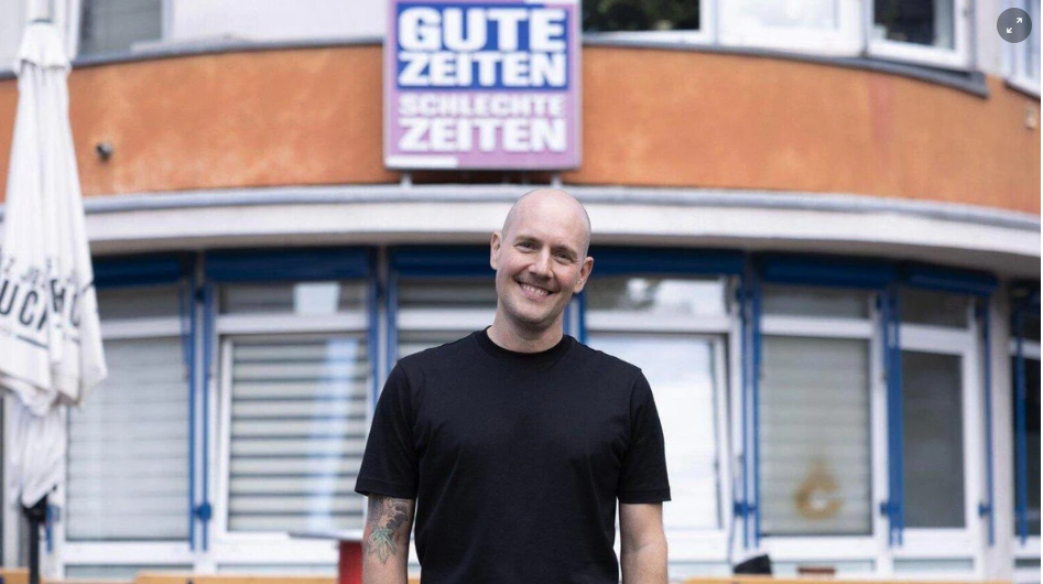 Ein lächelnder Mann steht vor einem Gebäude mit dem Schild „Gute Zeiten Schlechte Zeiten“.