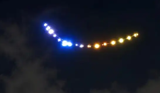 Leuchtende Punkte in Blau- und Gelbtönen ziehen eine Kurve am Nachthimmel.