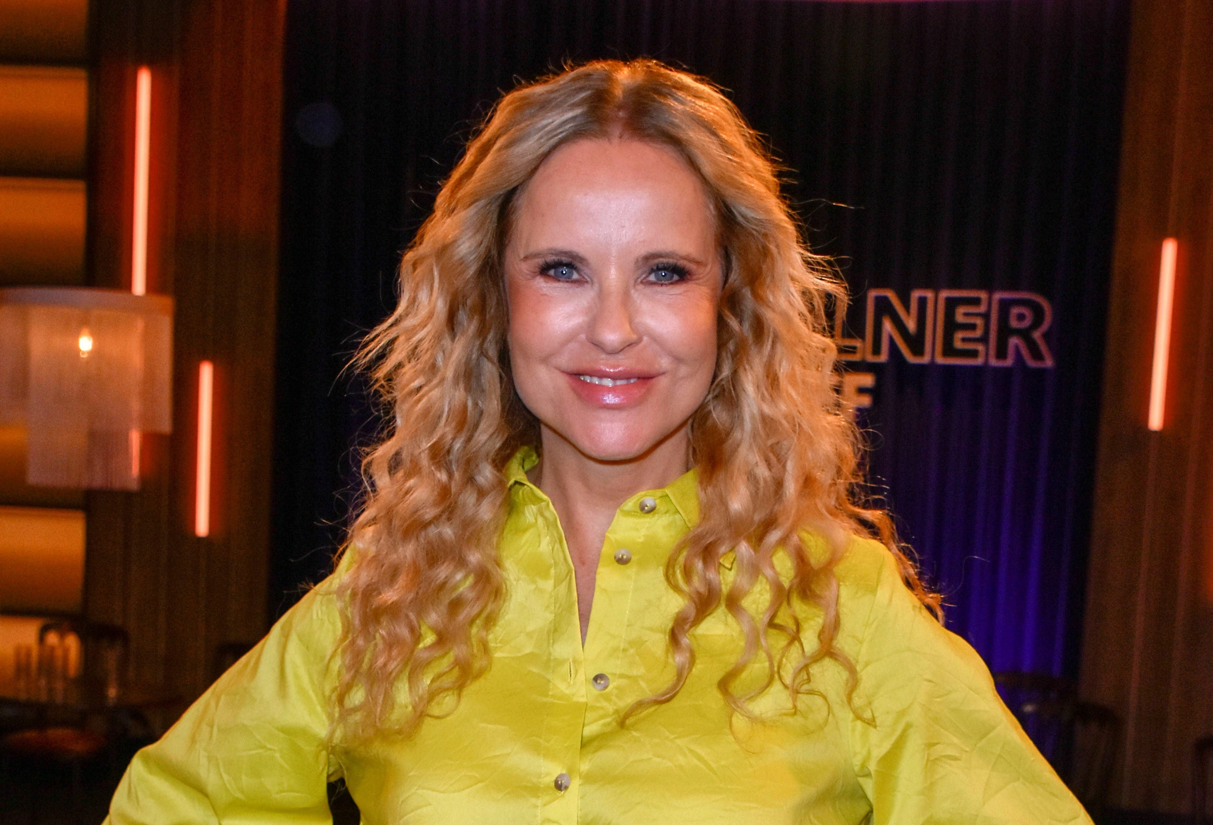 Frau mit blonden, lockigen Haaren und gelbem Hemd steht lächelnd in einem Studio.