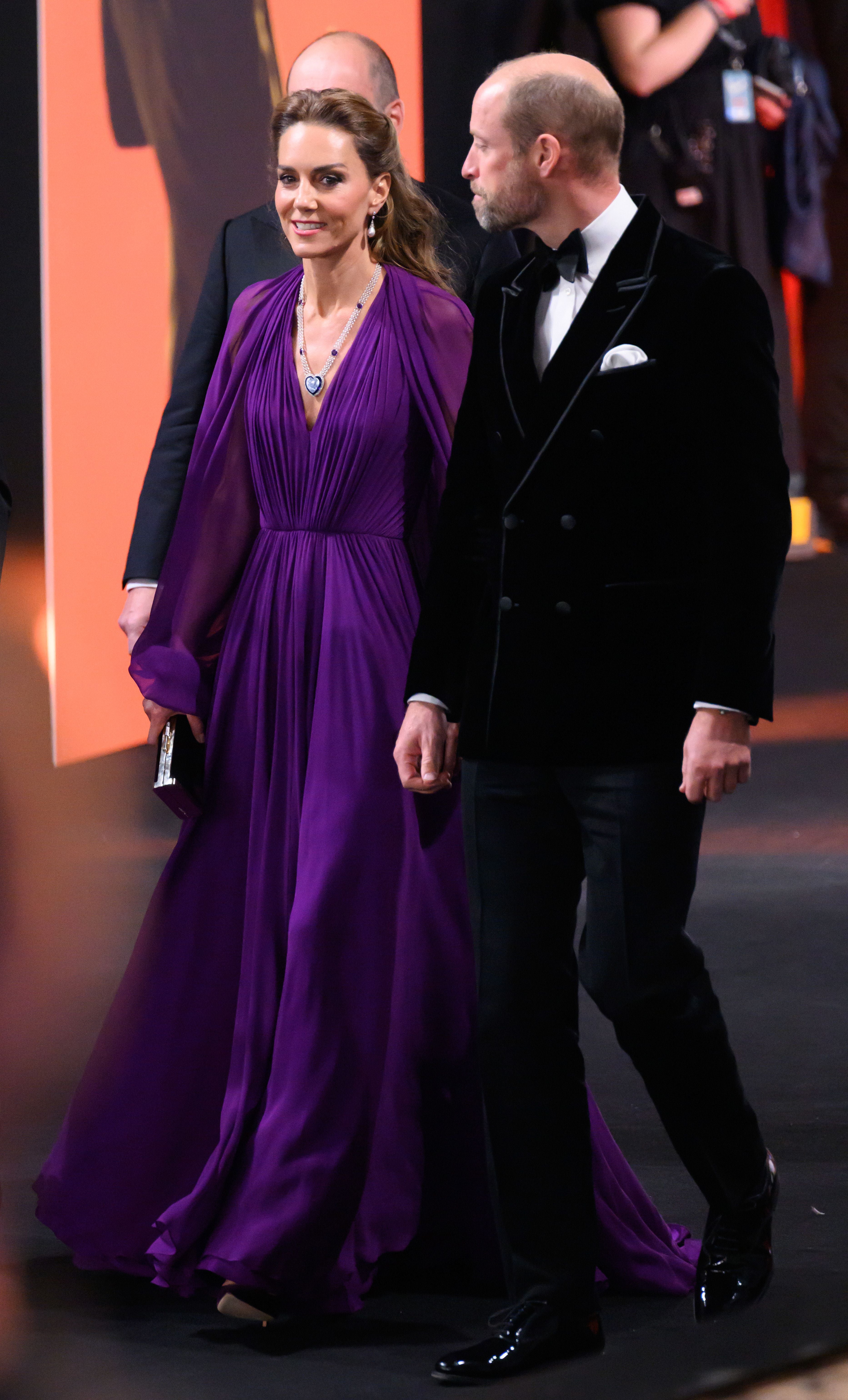 Eine Frau im lila Abendkleid und ein Mann im Smoking gehen gemeinsam über einen roten Teppich.