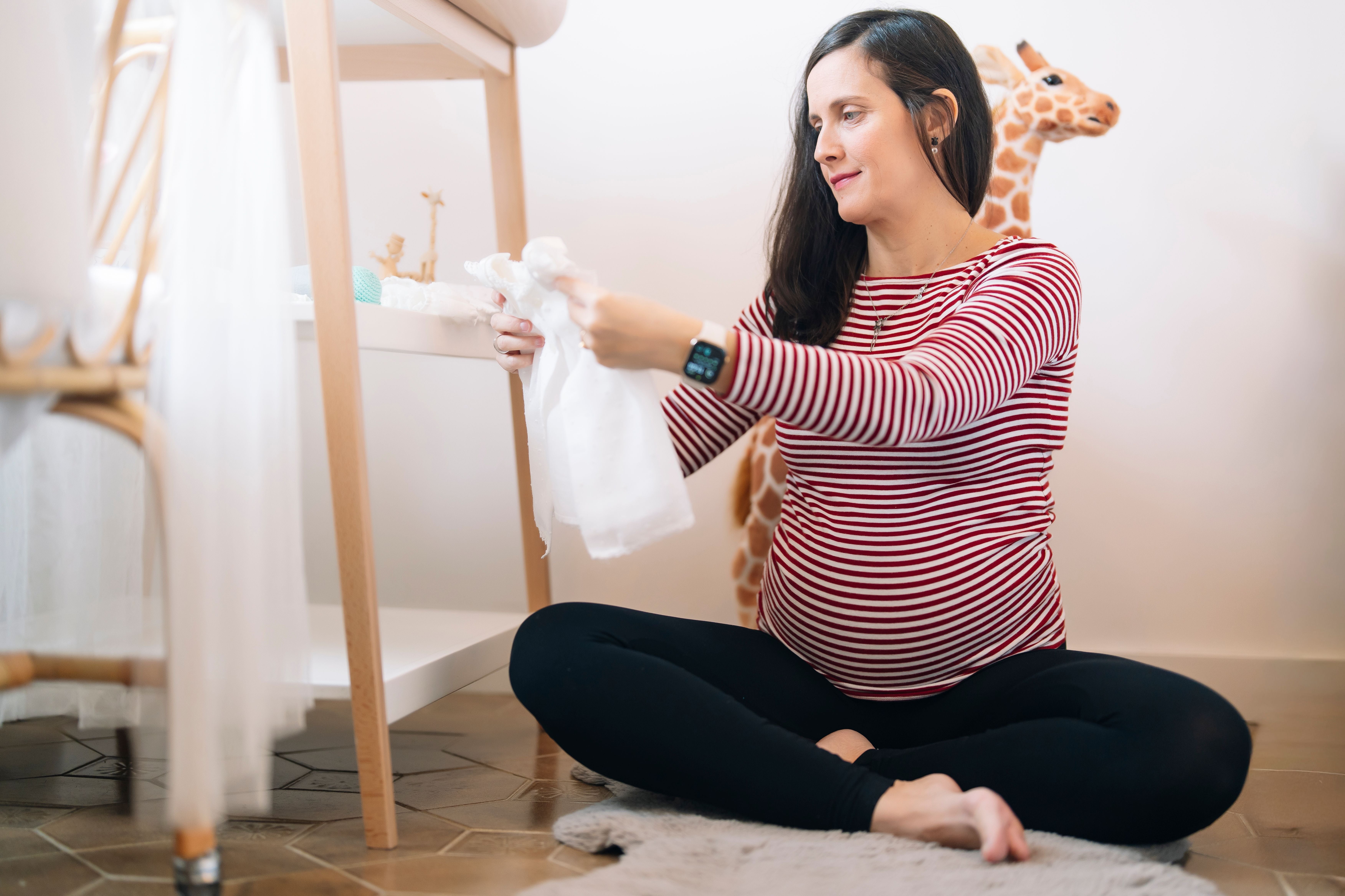 Schwangere Frau sitzt am Boden und sortiert Babykleidung im Kinderzimmer.