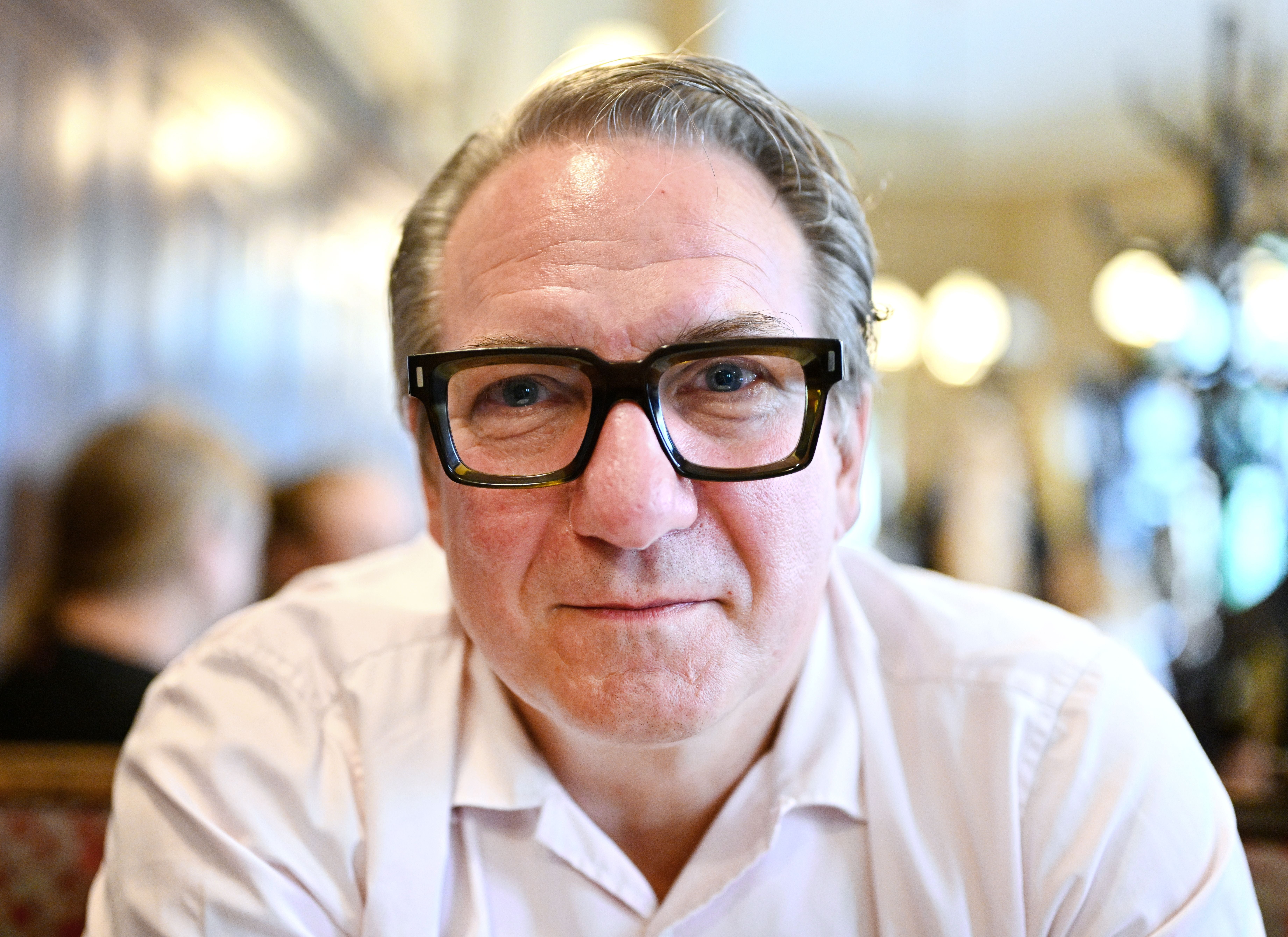 Mann mit Brille und weißem Hemd sitzt in einem Café und blickt in die Kamera.