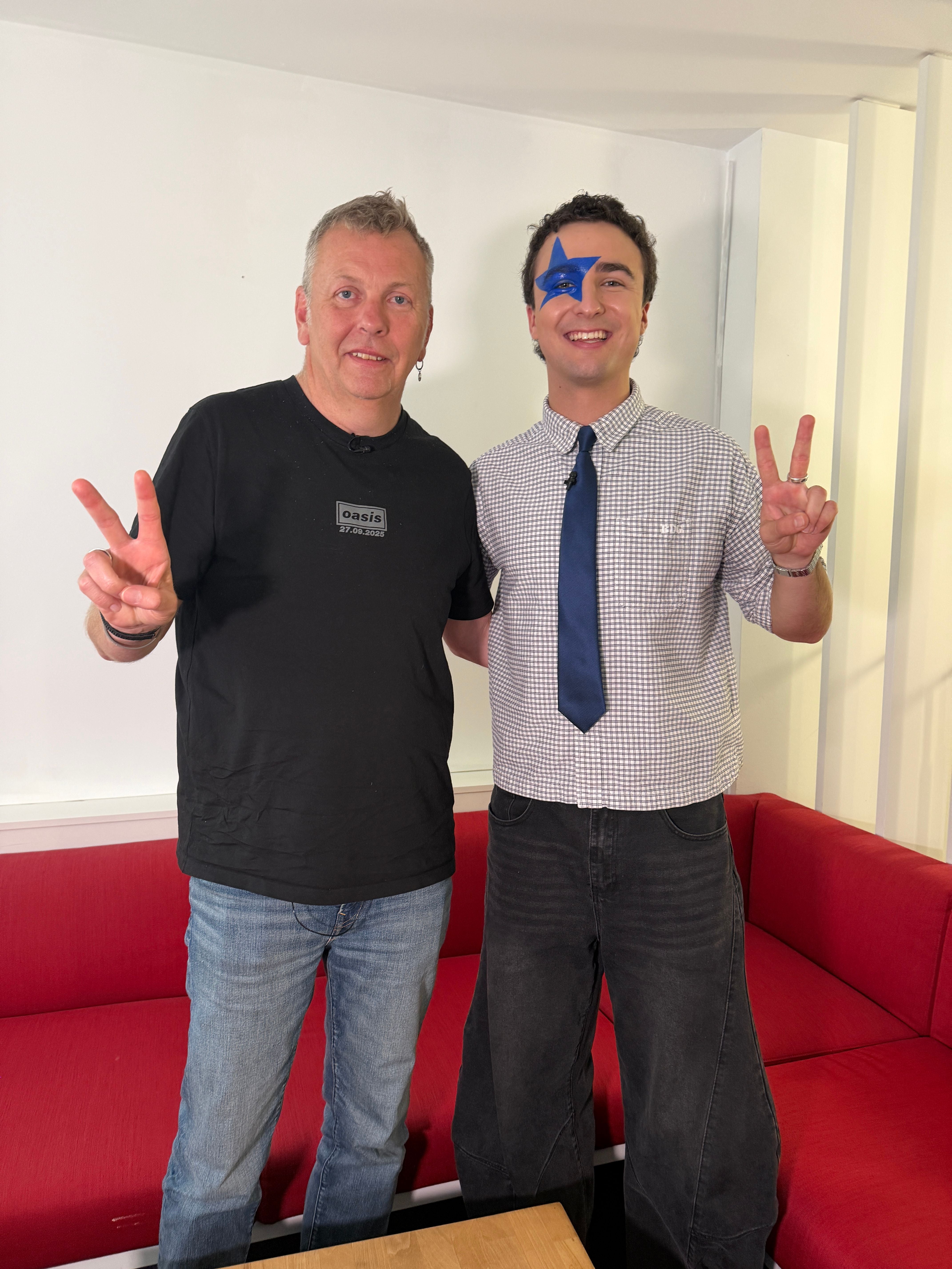 Zwei Männer stehen lächelnd auf einem roten Sofa und zeigen das Peace-Zeichen, einer mit blauem Stern im Gesicht.