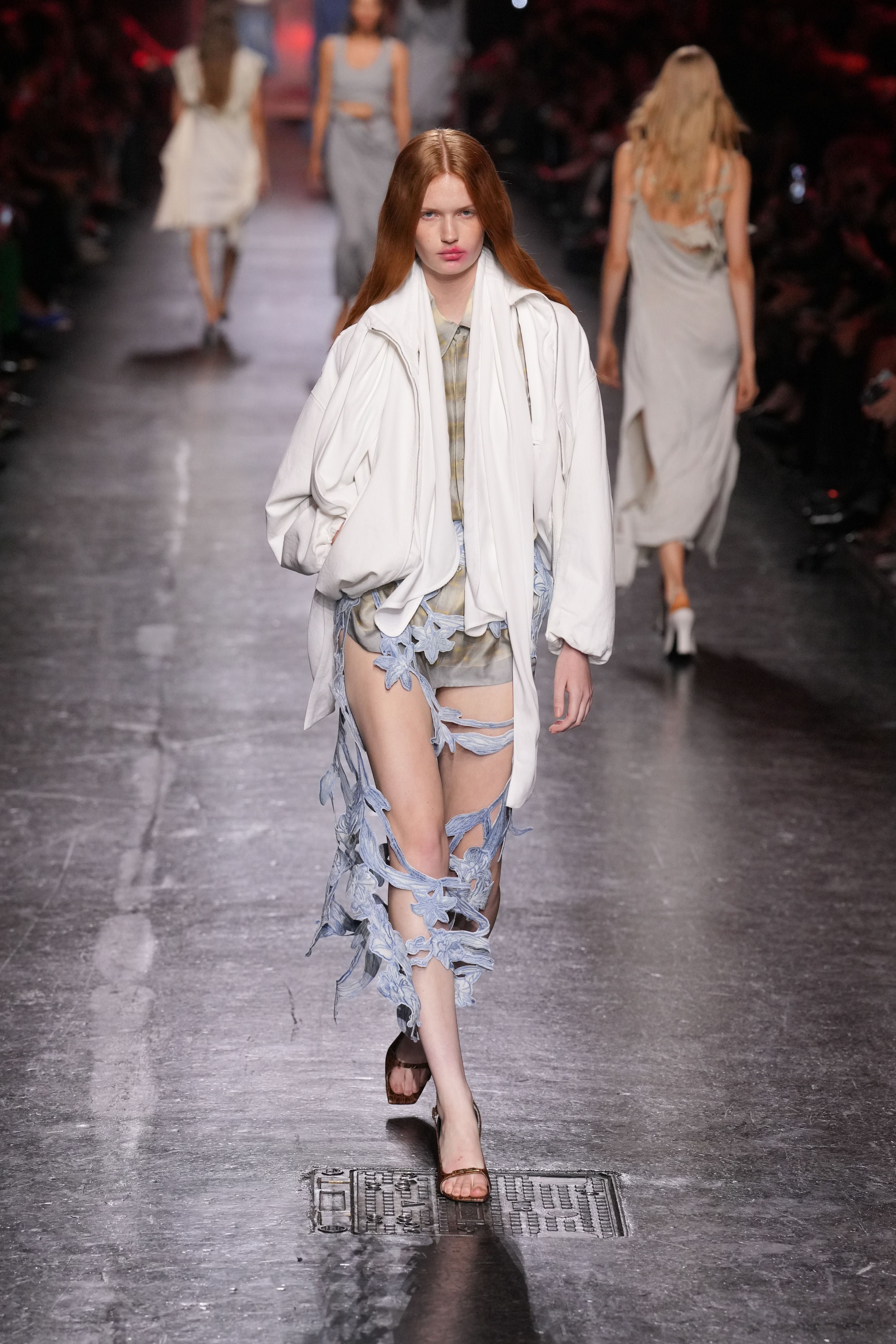 Model in weißer Jacke und Rock mit blauen Blütenapplikationen auf dem Laufsteg.