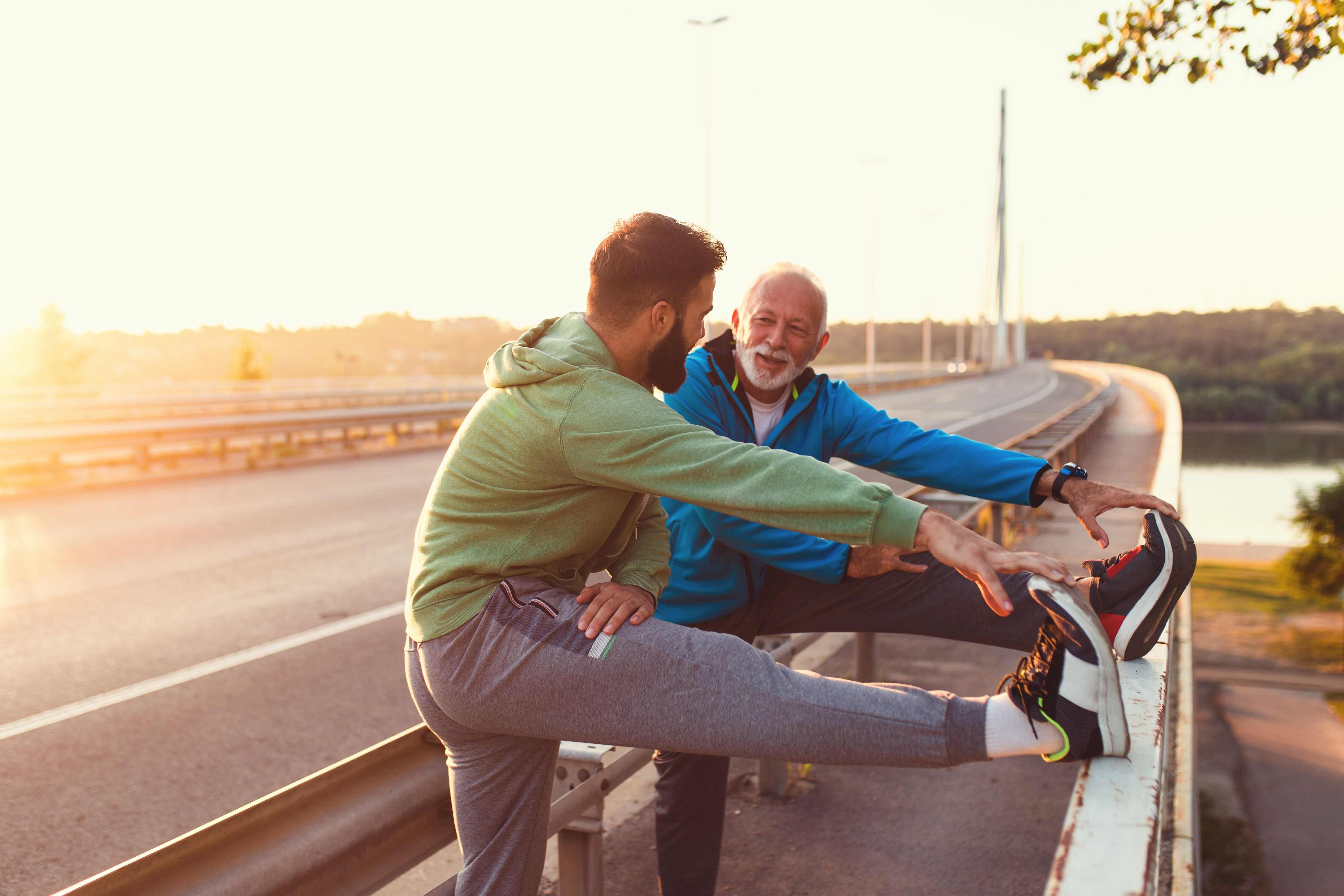Zwei Männer dehnen sich gemeinsam lachend auf einer Brücke im Sonnenlicht.