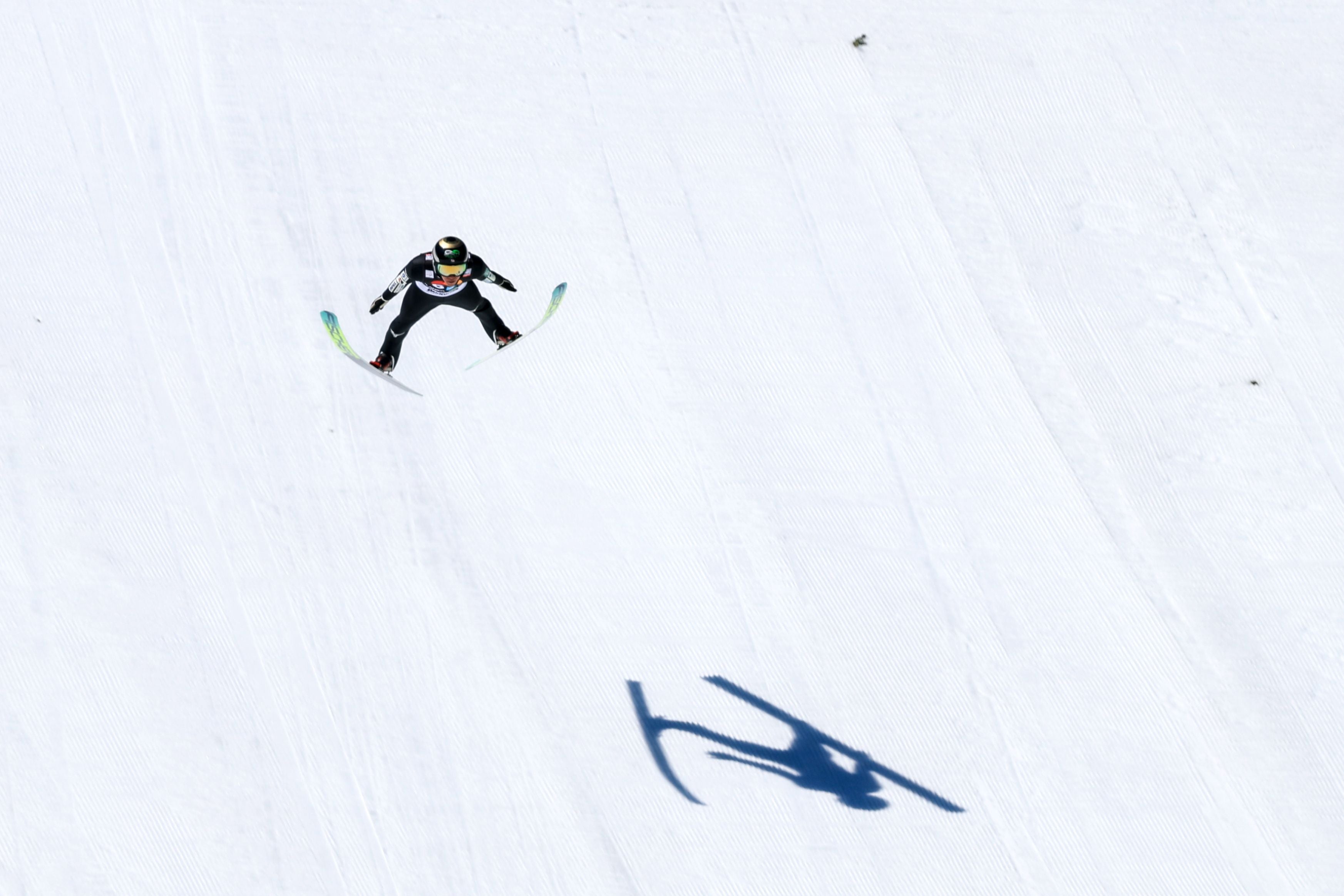 Ein Skispringer fliegt durch die Luft, sein Schatten ist deutlich auf dem Schnee zu sehen.