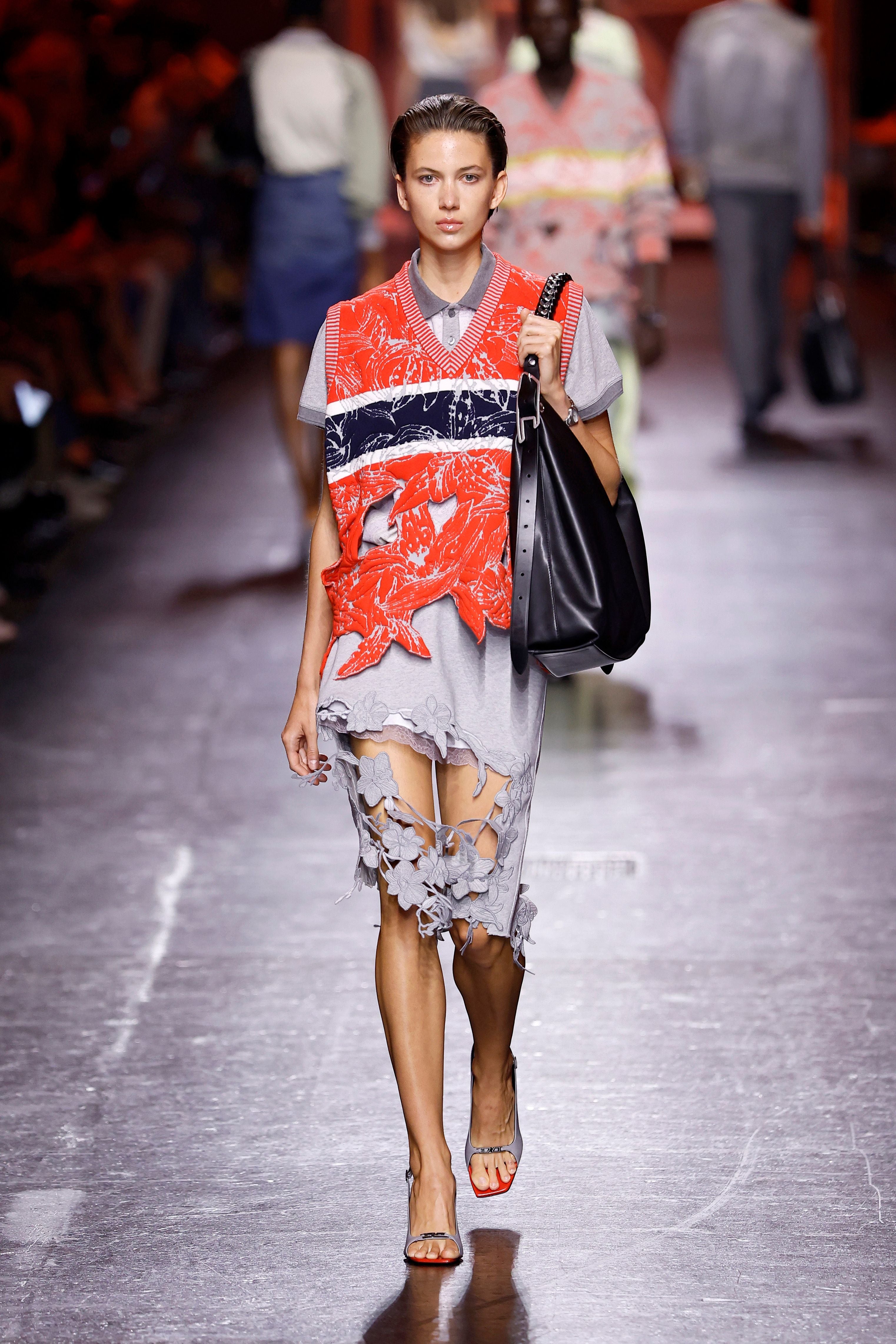 Model in grauem Kleid mit floralen Details und roter Weste auf dem Laufsteg einer Modenschau.