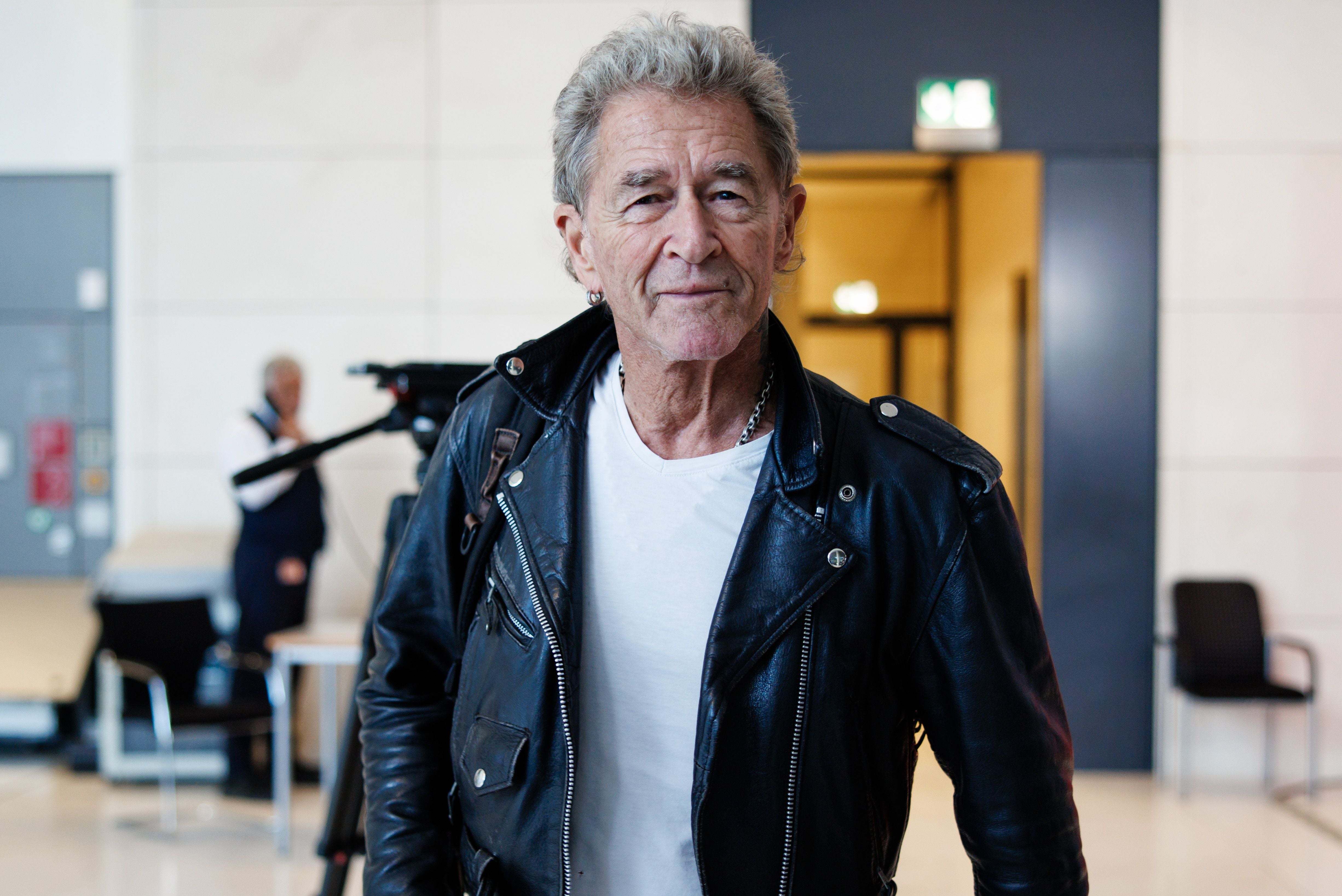 Ein älterer Mann in schwarzer Lederjacke steht in einem modernen, hellen Innenraum.