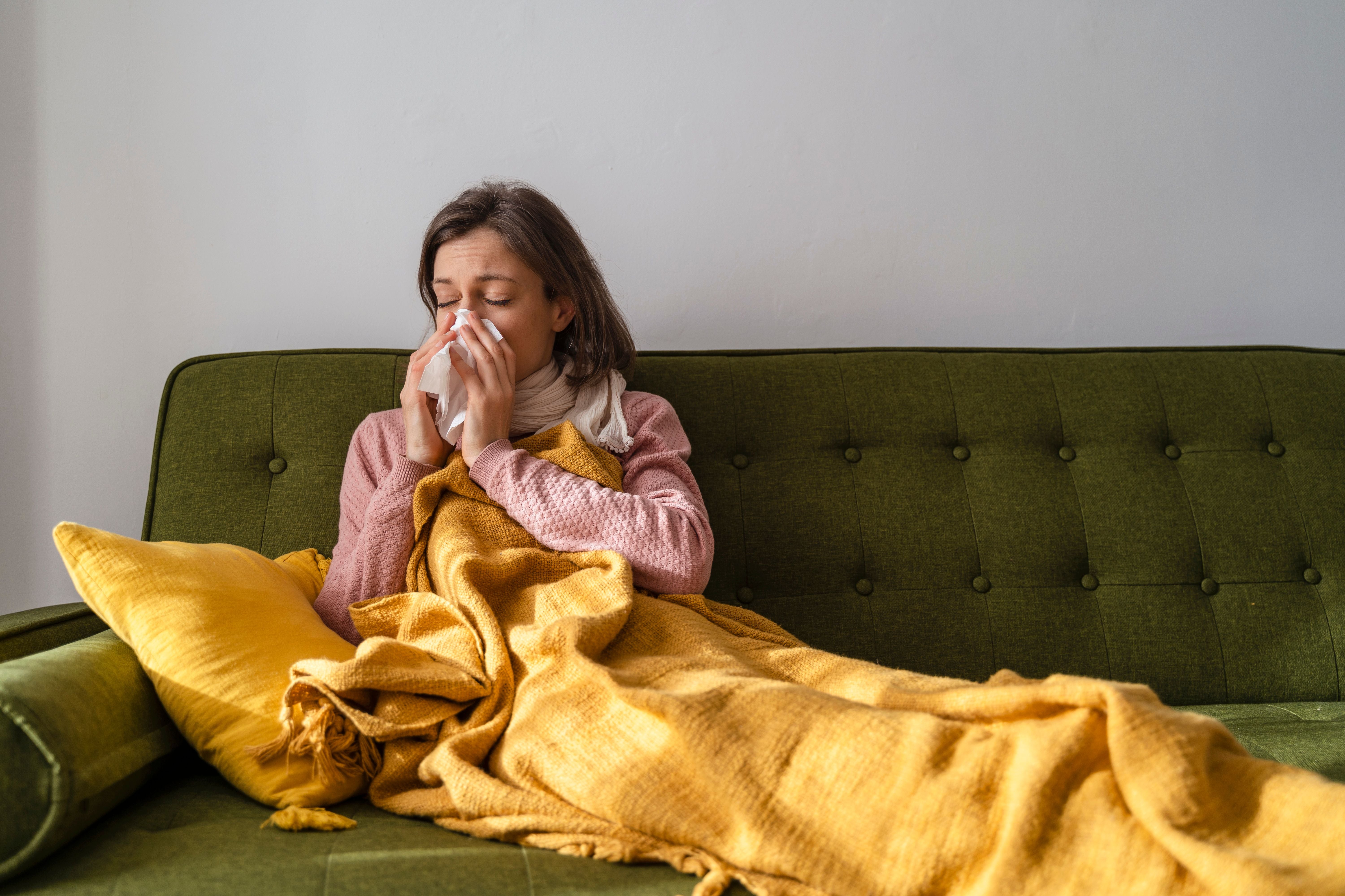 Frau mit Erkältung liegt mit Decke auf Sofa und putzt sich die Nase.