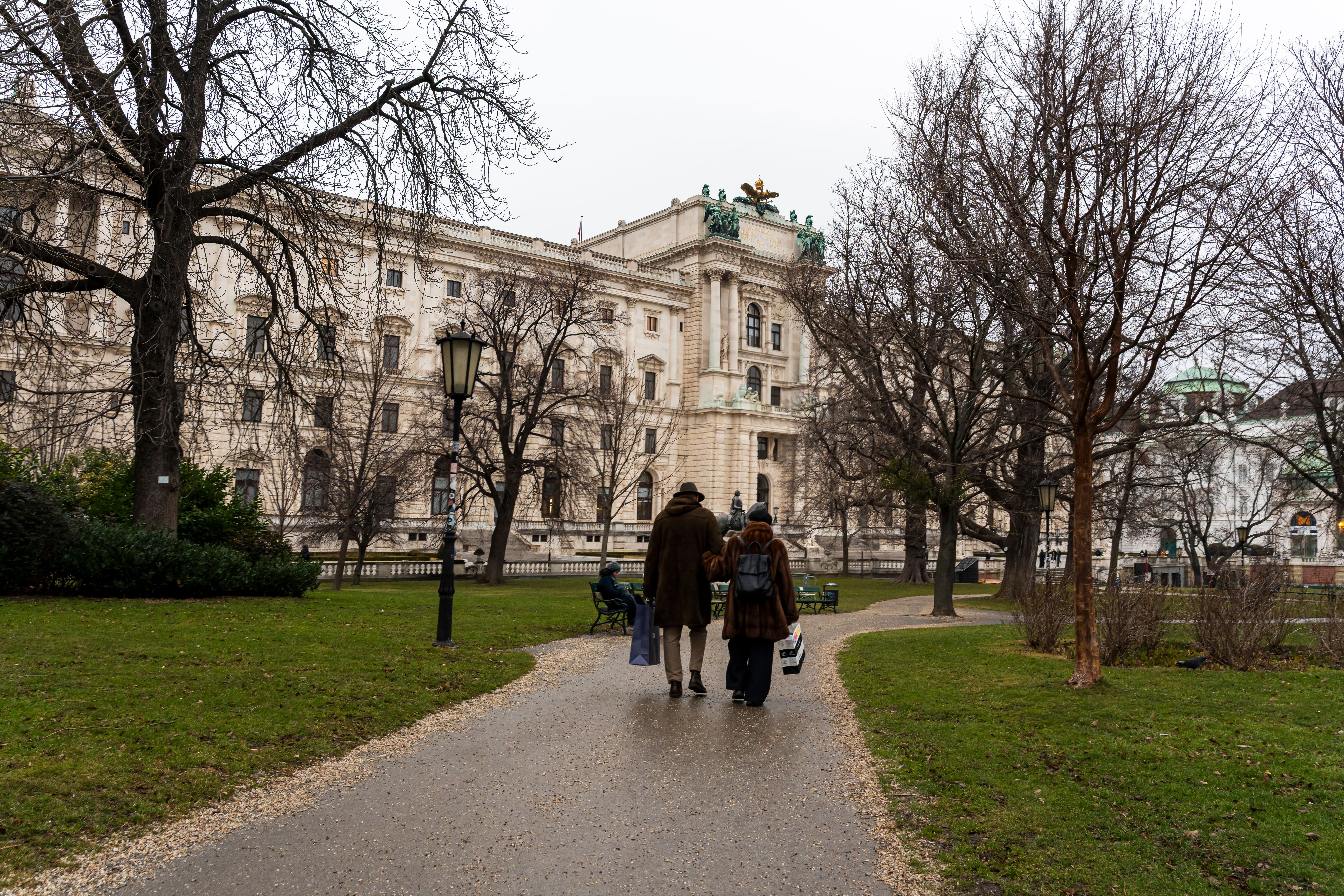 Drei Menschen spazieren auf einem Parkweg mit kahlen Bäumen und historischem Gebäude im Hintergrund.