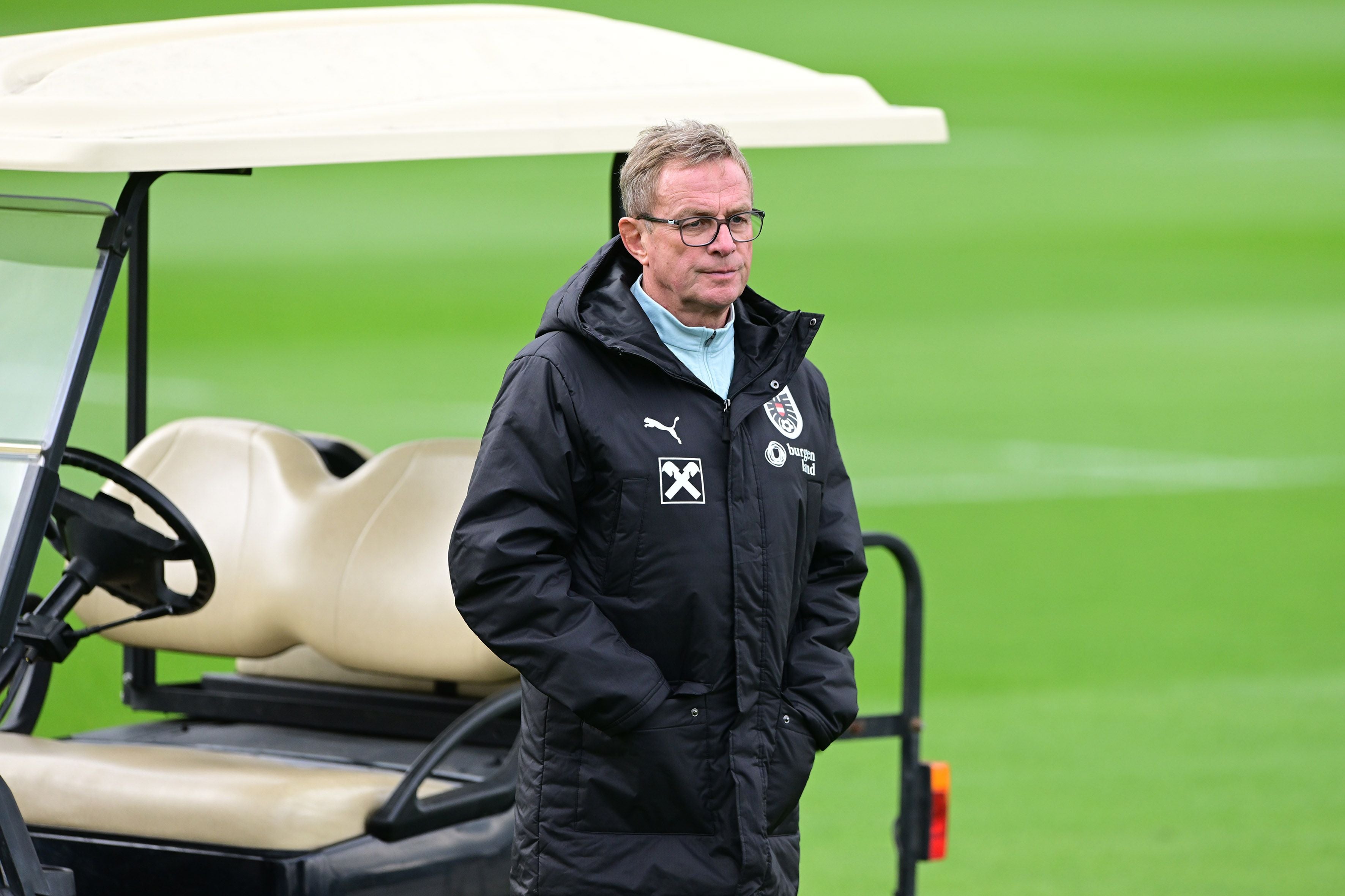 Ein Mann im schwarzen Trainingsanzug steht vor einem Golfcart auf einem Fußballfeld.