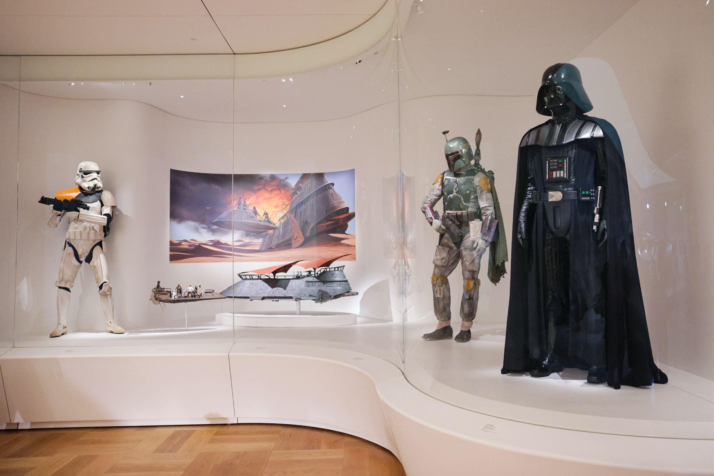 Drei Star Wars Kostüme und Raumschiff-Modelle in einer Museumsvitrine ausgestellt.