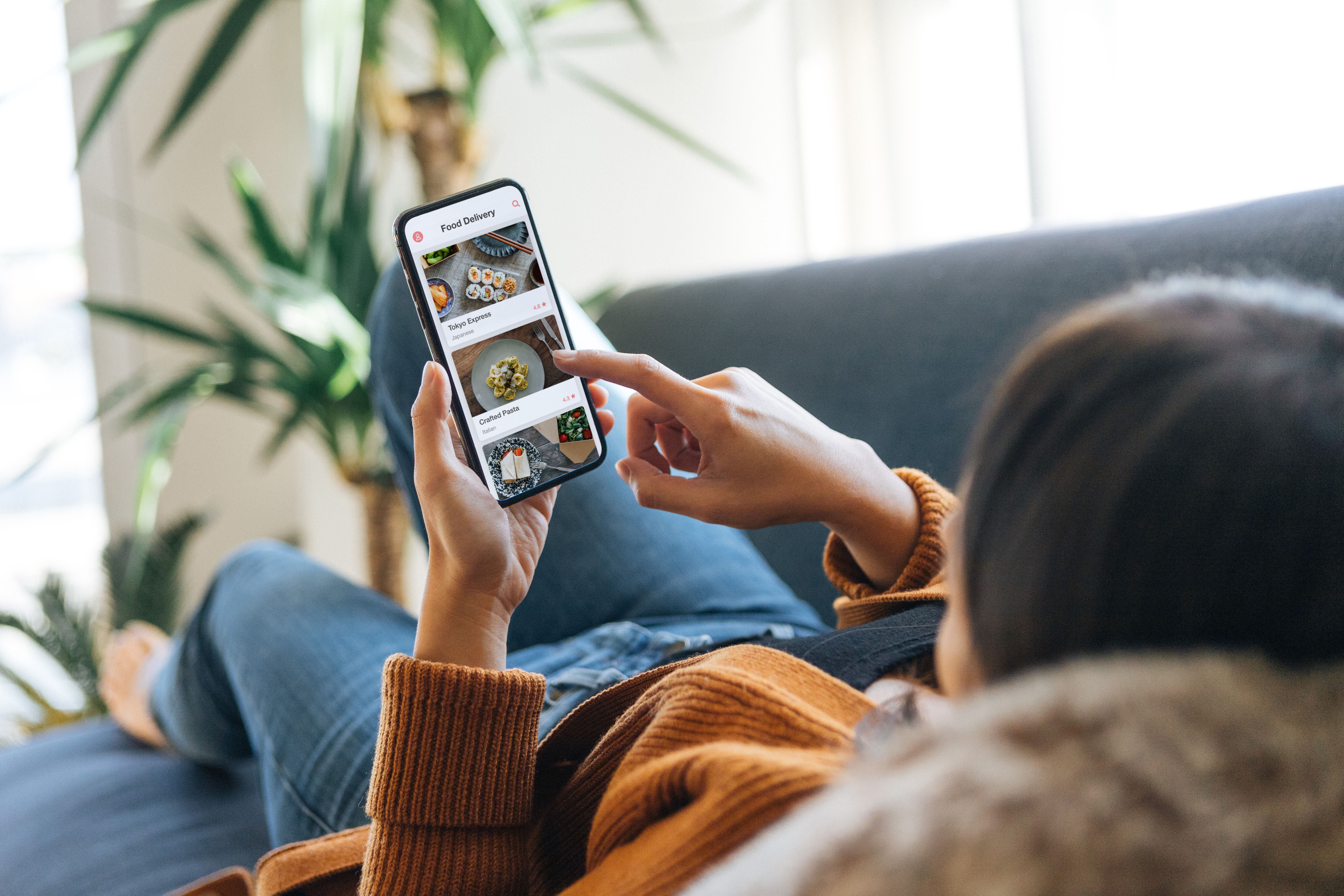 Junge Frau liegt auf dem Sofa und bestellt Essen per App auf ihrem Smartphone.