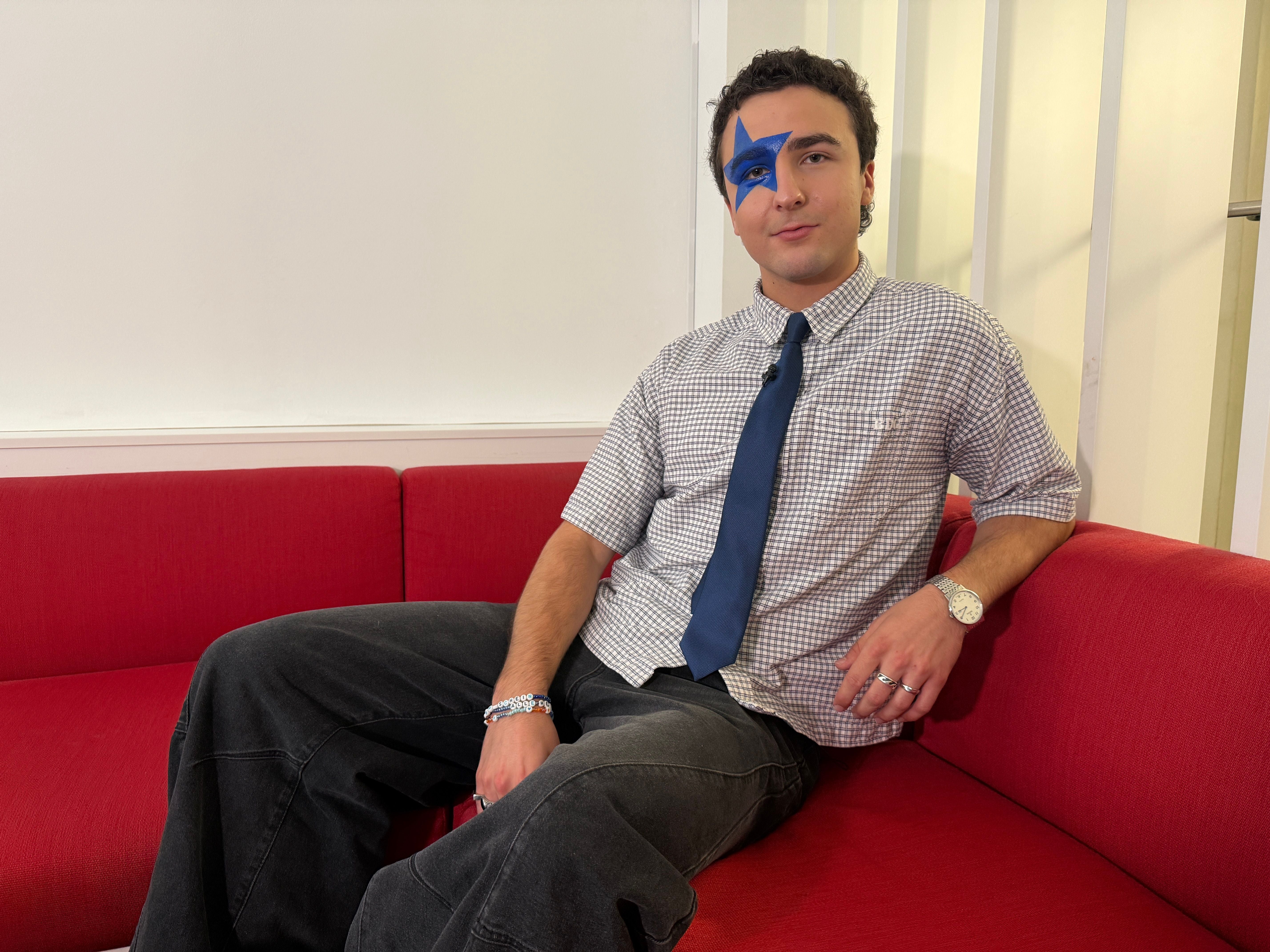 Junger Mann mit blauem Stern im Gesicht sitzt auf rotem Sofa, trägt Hemd und Krawatte.
