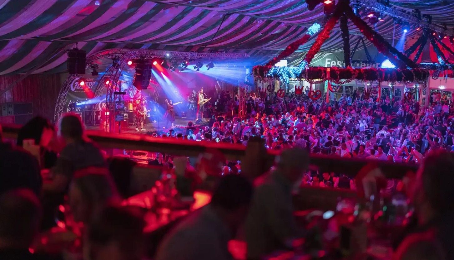 Blick auf ein bunt beleuchtetes Festzelt mit Bühne und tanzender Menschenmenge auf der Kaiser Wiesn.