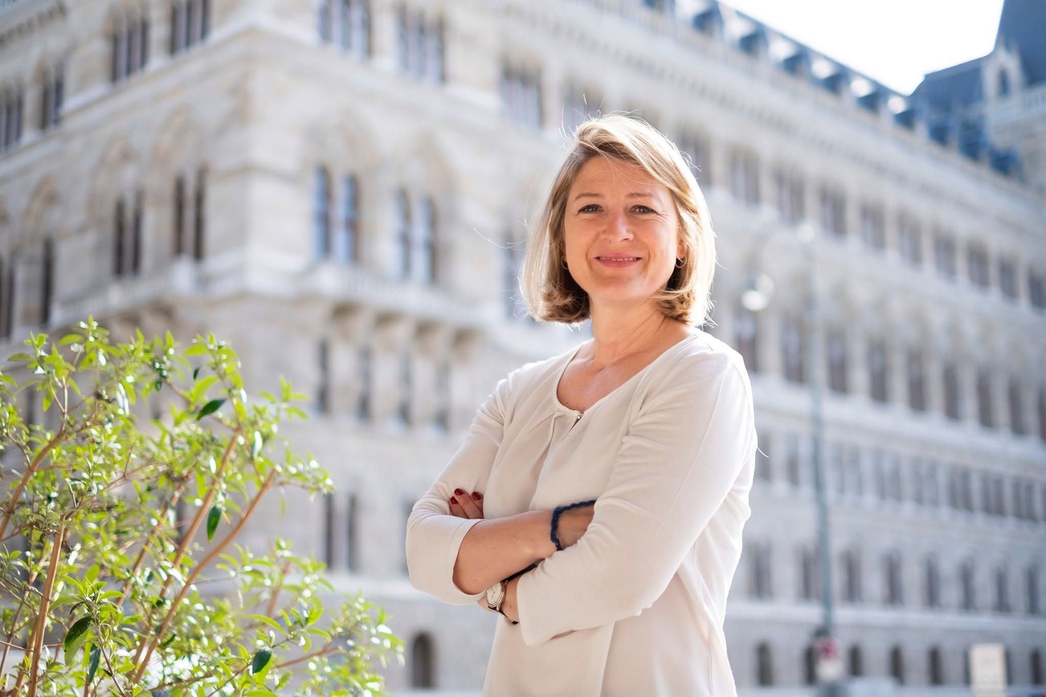 Eine Frau steht lächelnd mit verschränkten Armen vor einem historischen Gebäude in der Stadt.