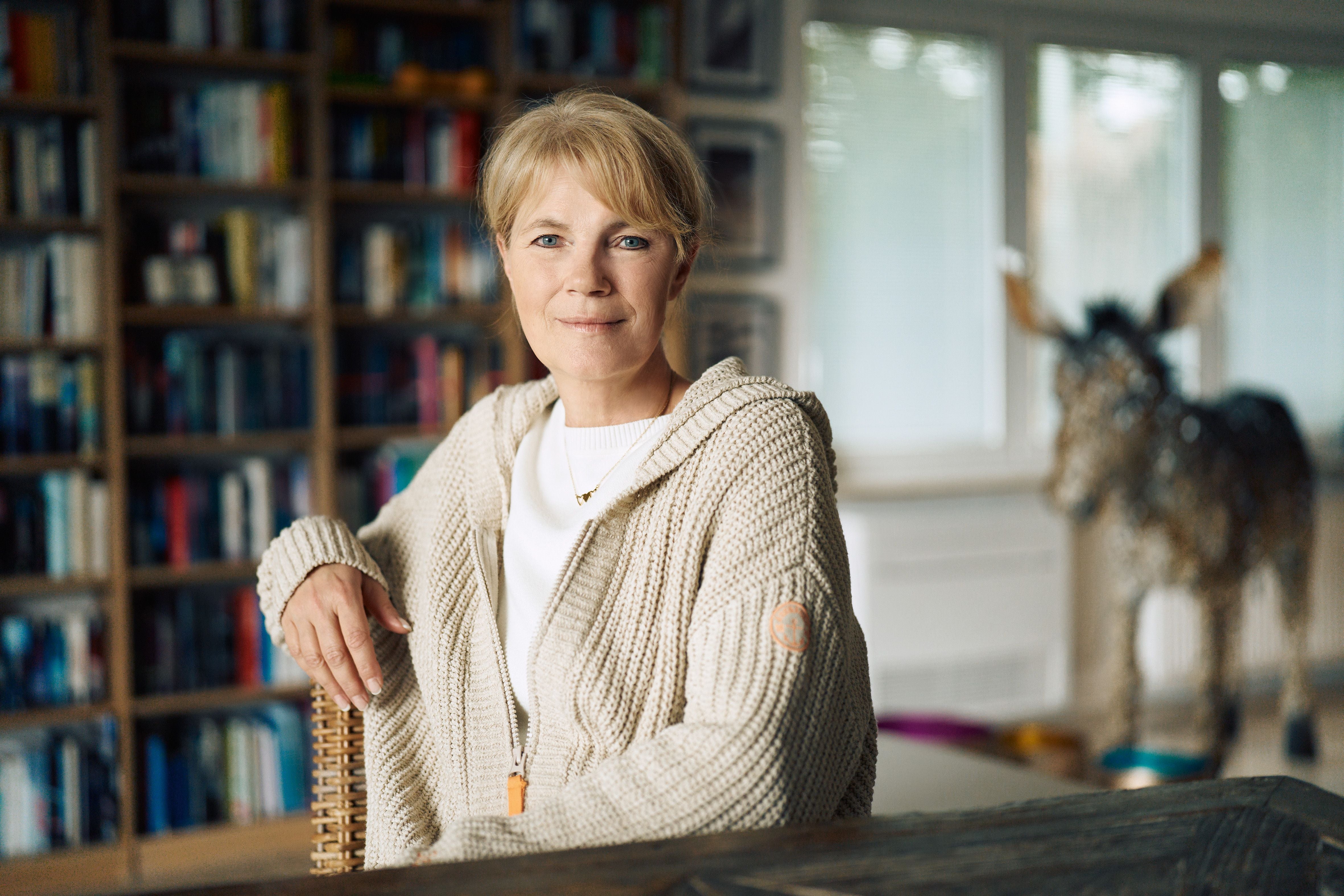 Frau mit blonder Kurzhaarfrisur sitzt vor einem Bücherregal in einem hellen Raum.