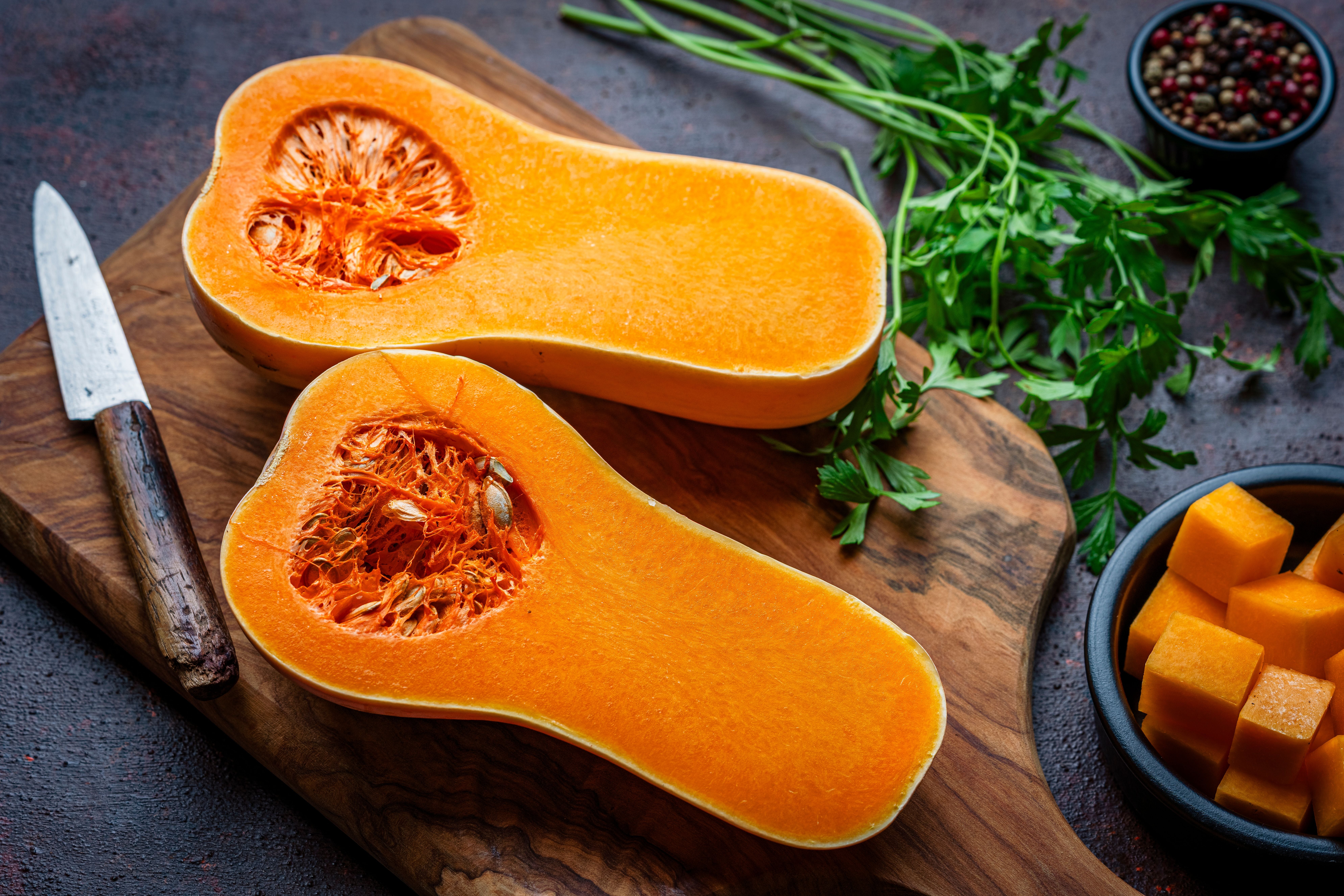 Halbierter Butternut-Kürbis auf einem Holzbrett mit Kräutern, Messer und Gewürzen.
