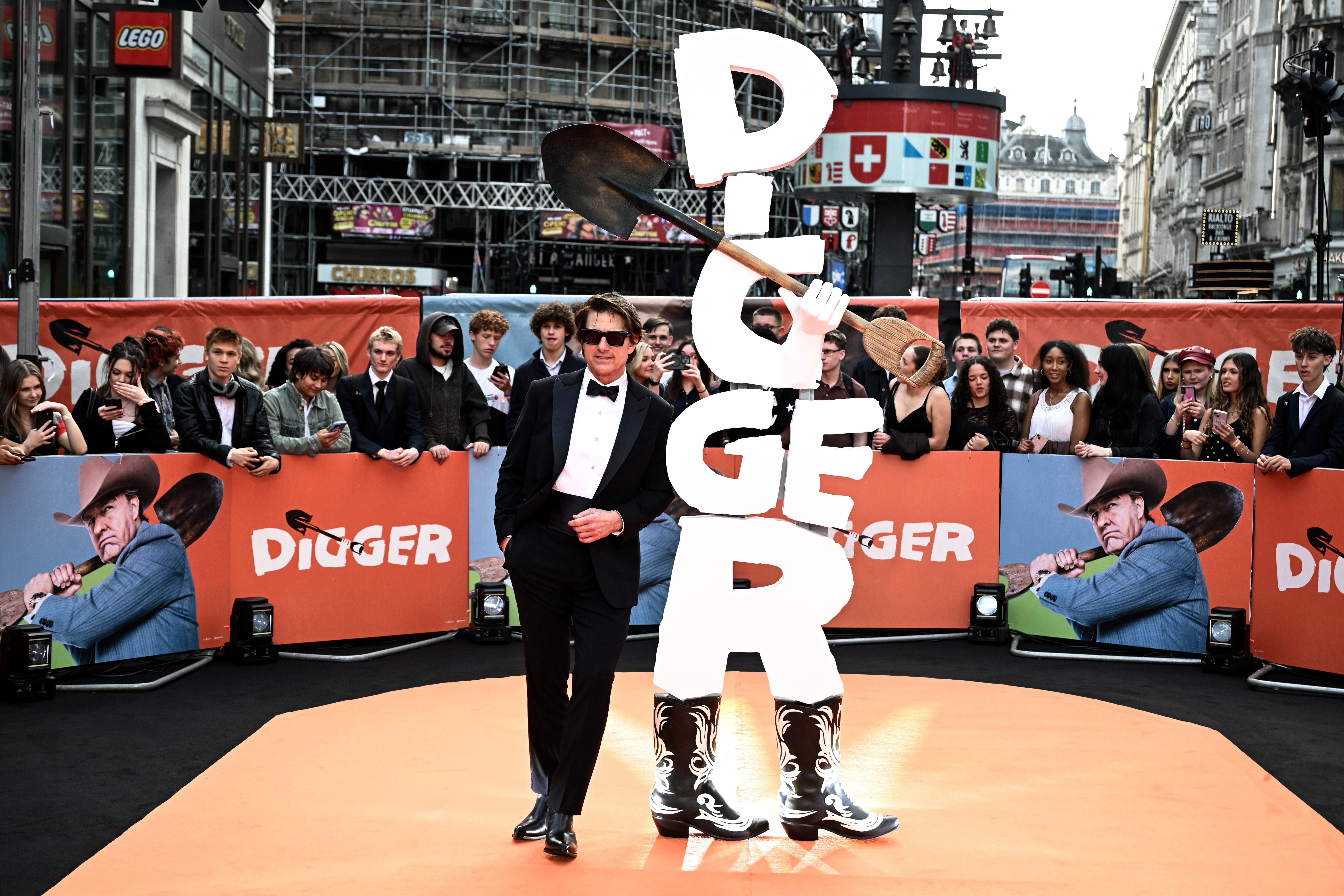 Ein Mann im Anzug steht lässig vor einem großen "DIGGER"-Schild mit Schaufel auf rotem Teppich.