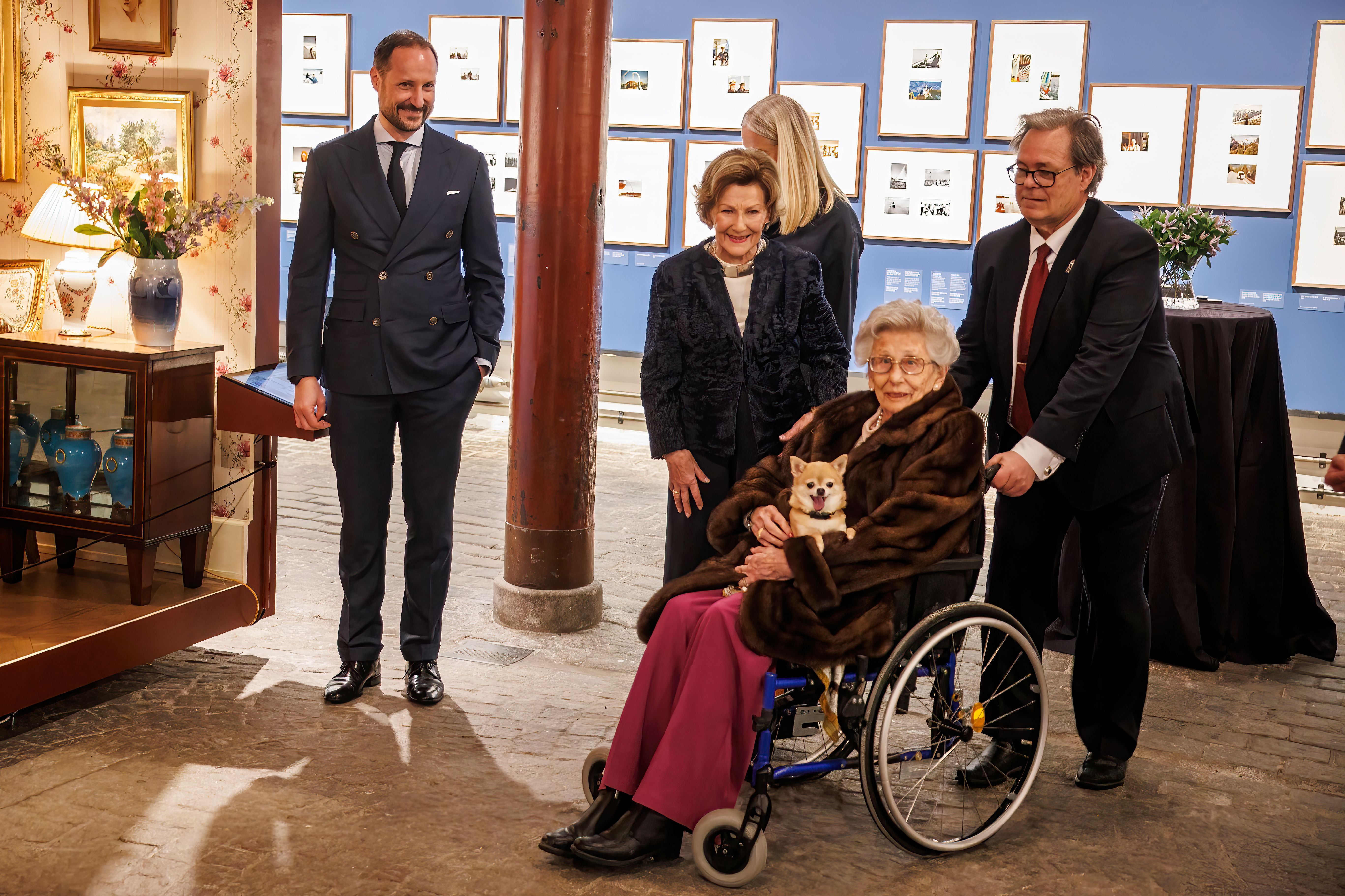 Mehrere festlich gekleidete Personen, eine Frau im Rollstuhl mit kleinem Hund, bei einer Kunstausstellung.