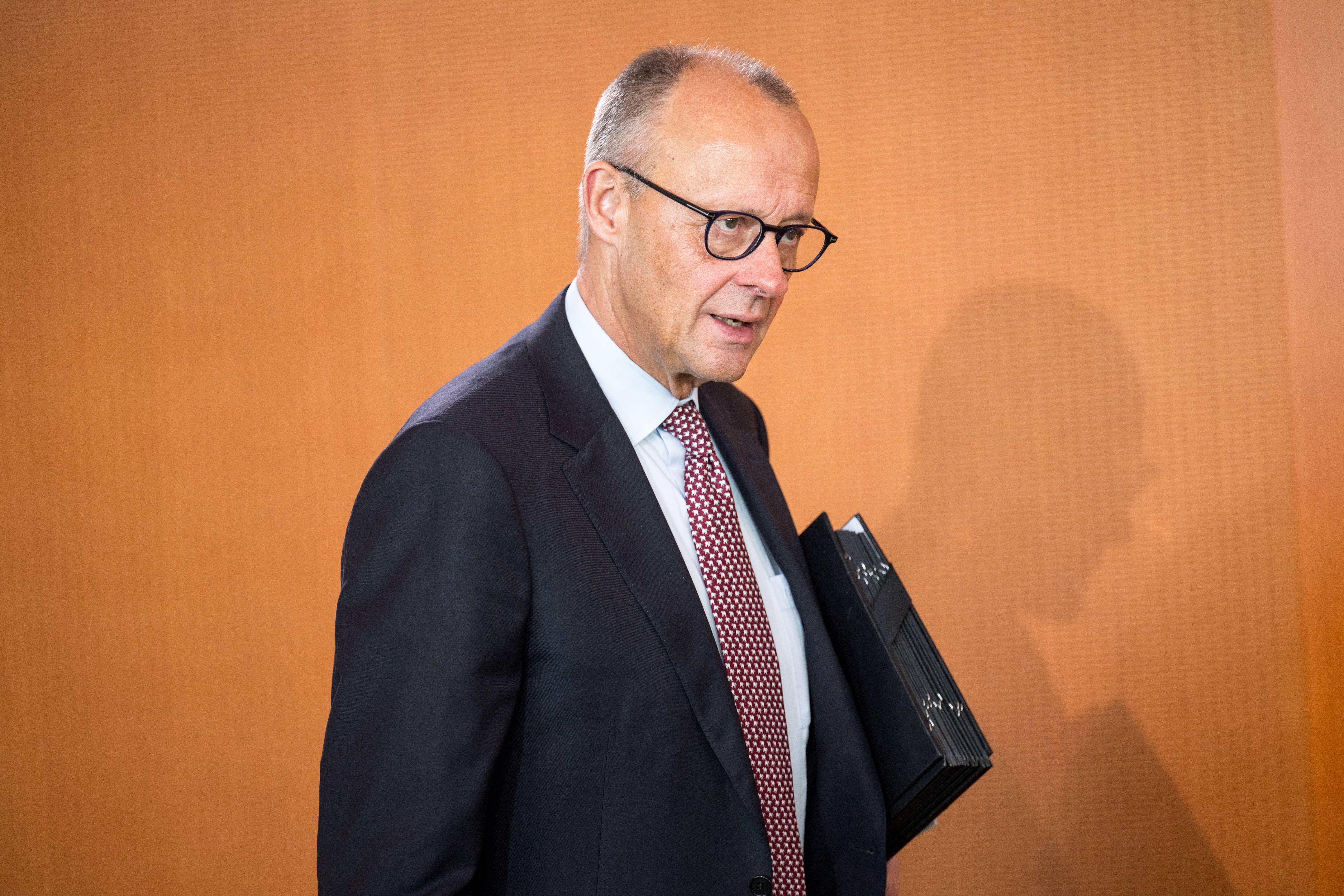 Ein Mann im Anzug mit Brille hält Aktenordner und steht vor einer orangefarbenen Wand.