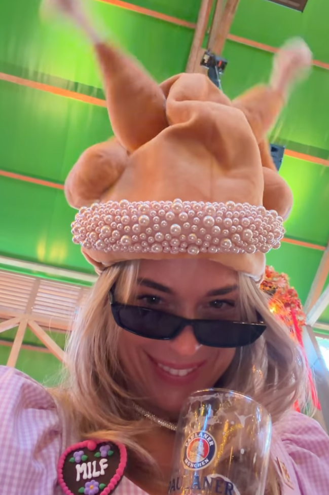 Frau mit Truthahn-Hut, Sonnenbrille und Bierglas auf einem Fest, trägt rosa Dirndl.