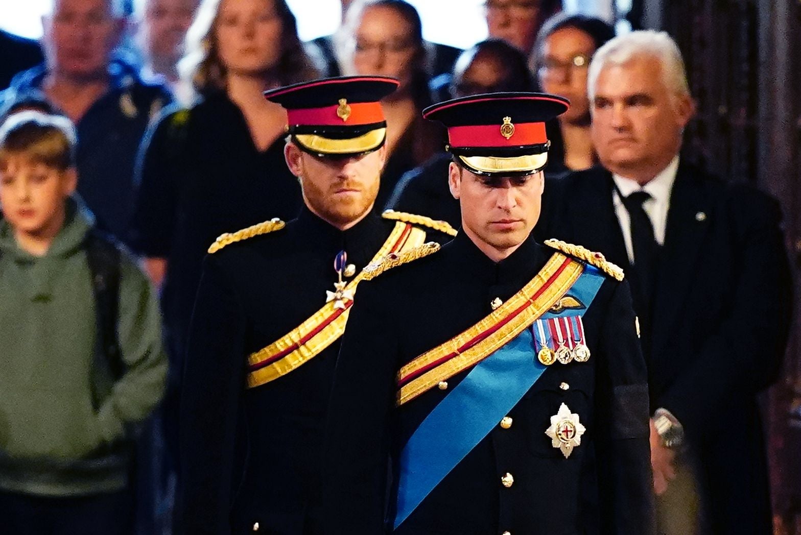 Zwei Männer in Militäruniform mit Schärpen und Mützen stehen ernst nebeneinander, Menschen im Hintergrund.