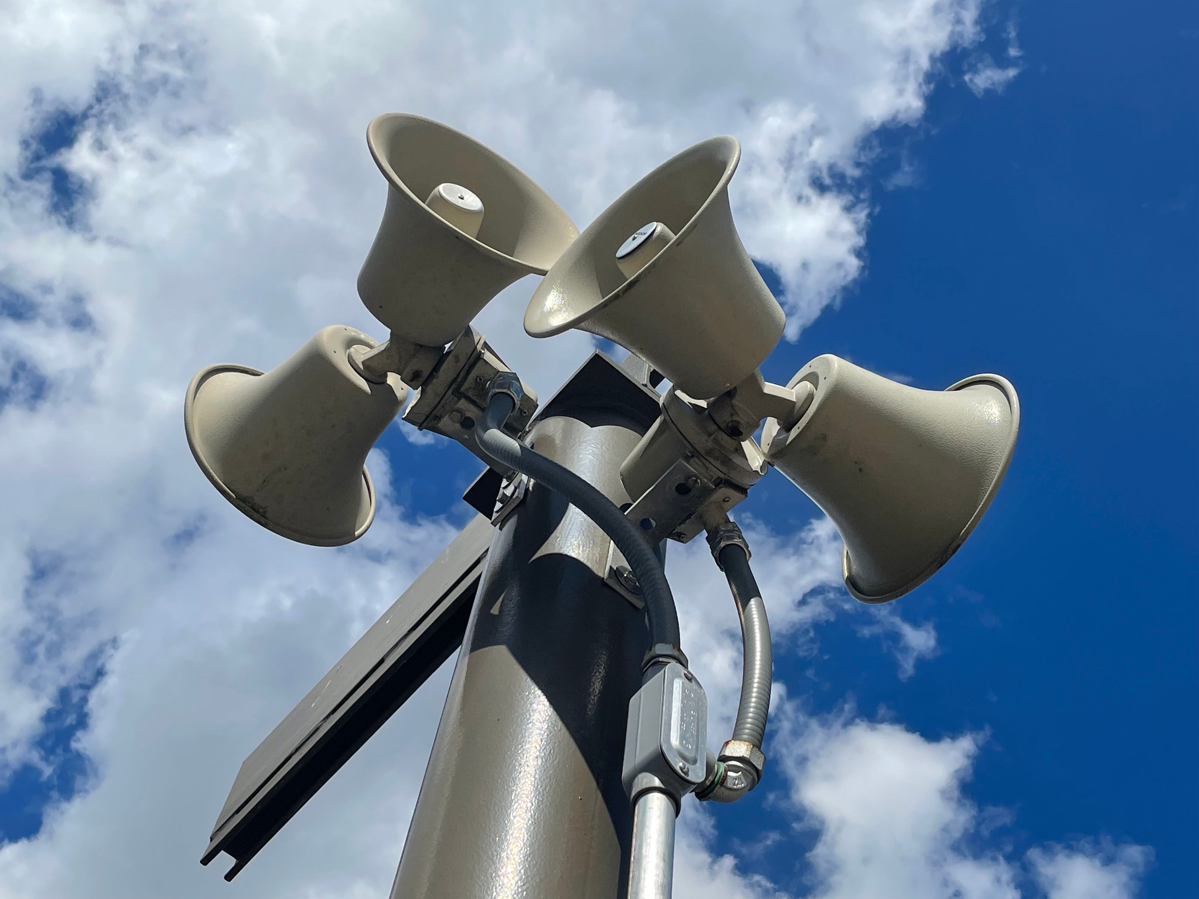 Vier Lautsprecher an einem Mast vor blauem Himmel mit weißen Wolken.