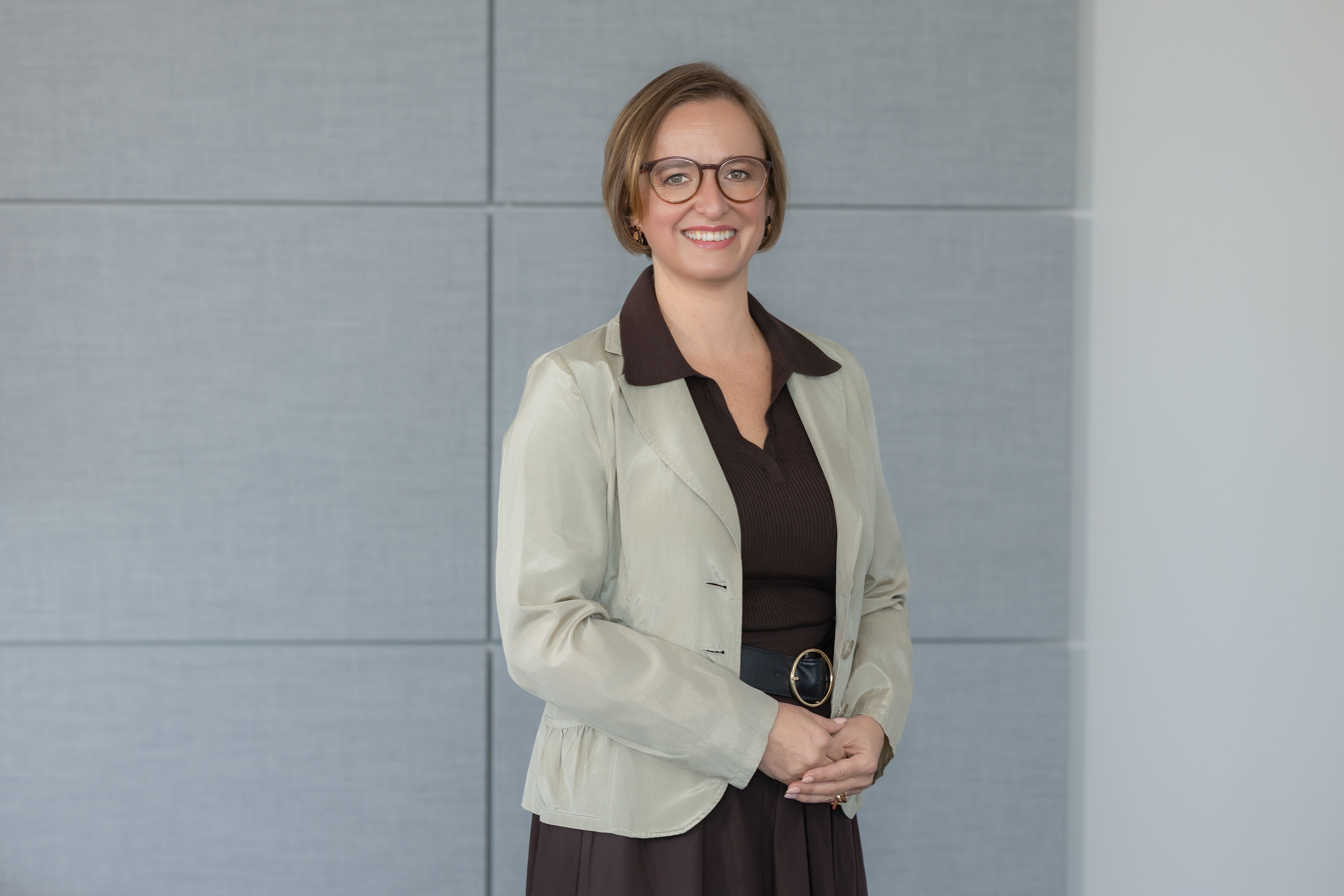 Frau mit Brille, hellem Blazer und dunklem Oberteil lächelt vor grauem Hintergrund in die Kamera.