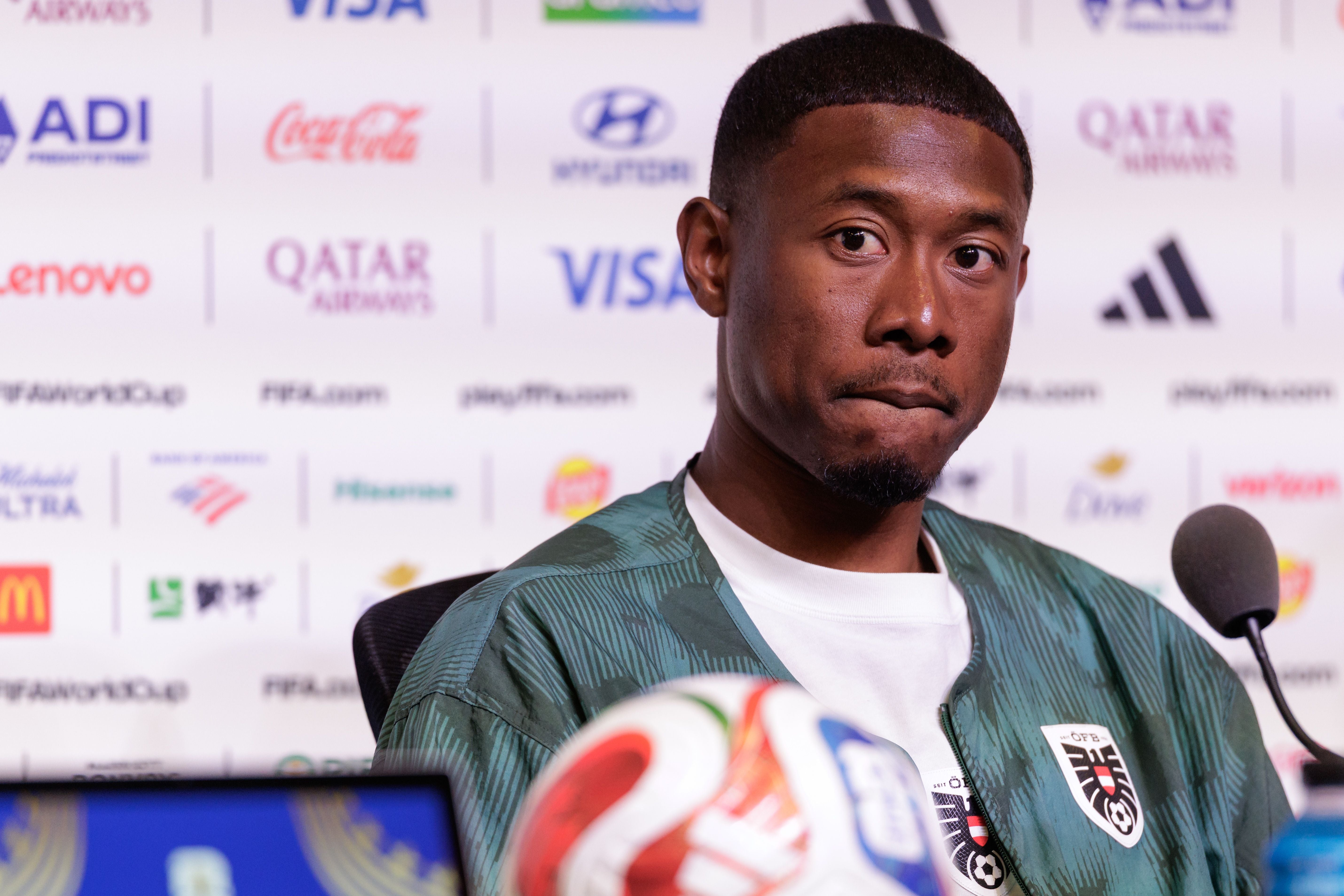 Ein Fußballspieler im Trainingsanzug spricht auf einer Pressekonferenz vor FIFA-Werbetafeln.
