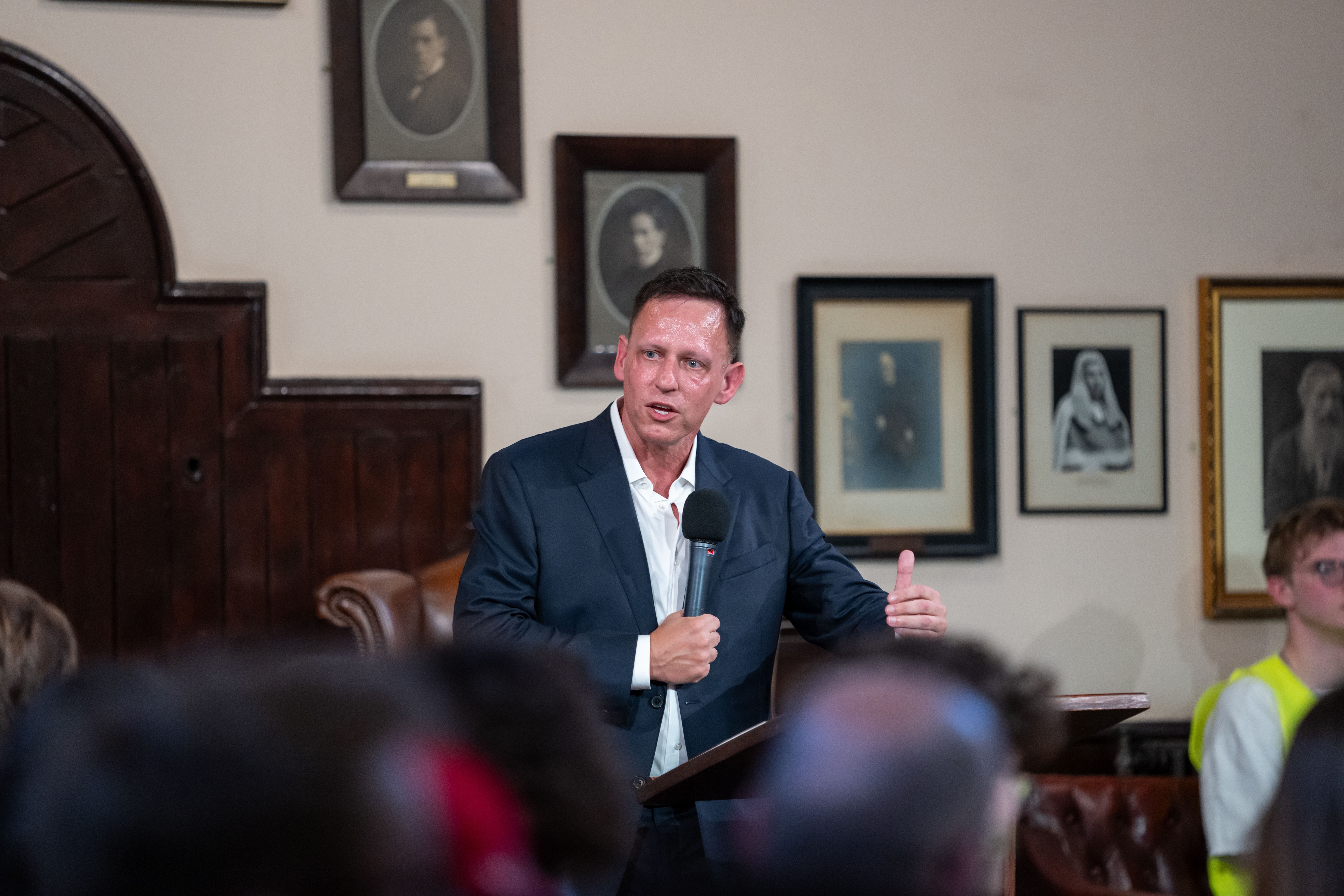 Ein Mann im Anzug spricht mit Mikrofon vor Publikum in einem Saal mit gerahmten Fotos an der Wand.