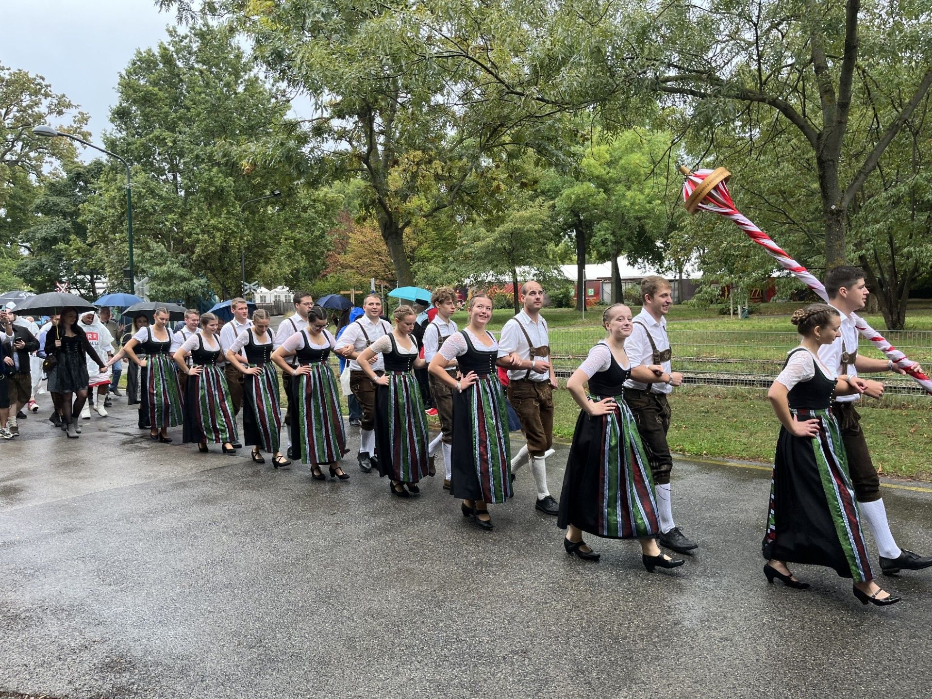 Gruppe in traditioneller Tracht marschiert beim Festzug auf nasser Straße, Bäume im Hintergrund.