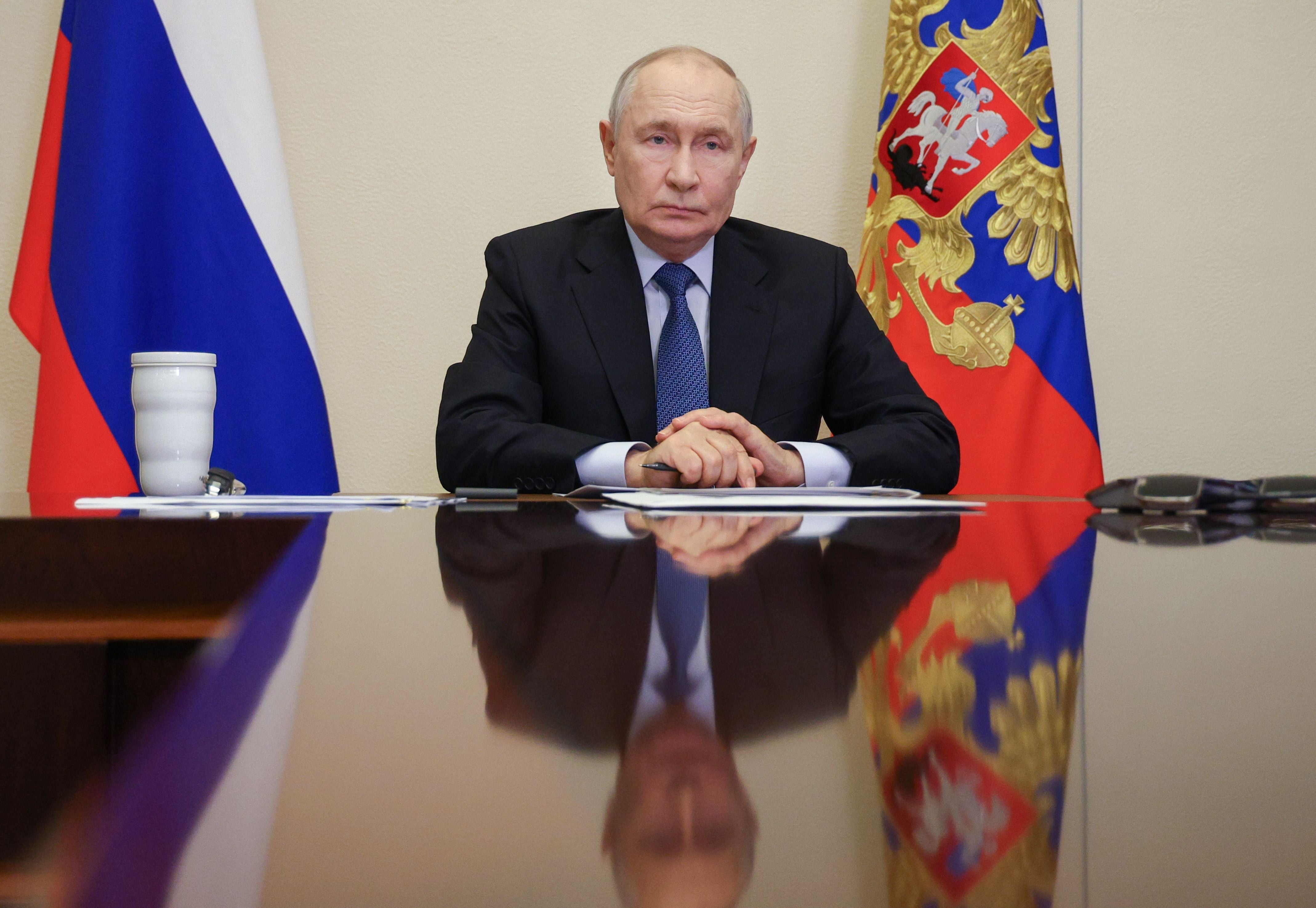Ein Mann im Anzug sitzt an einem Tisch mit russischen Flaggen im Hintergrund.