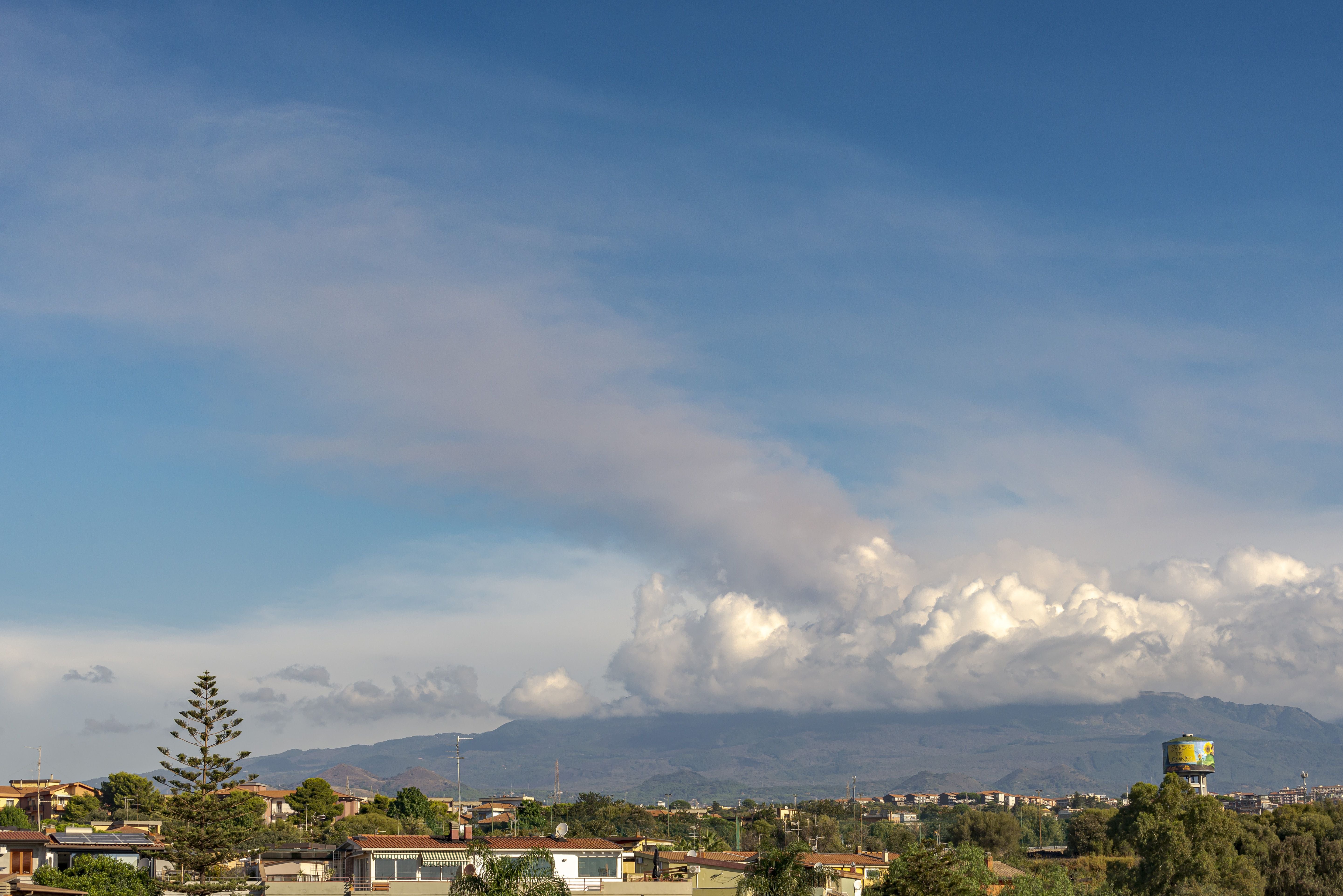 Vulkanische Wolken steigen vom Ätna auf, im Vordergrund eine Stadtlandschaft bei klarem Himmel.