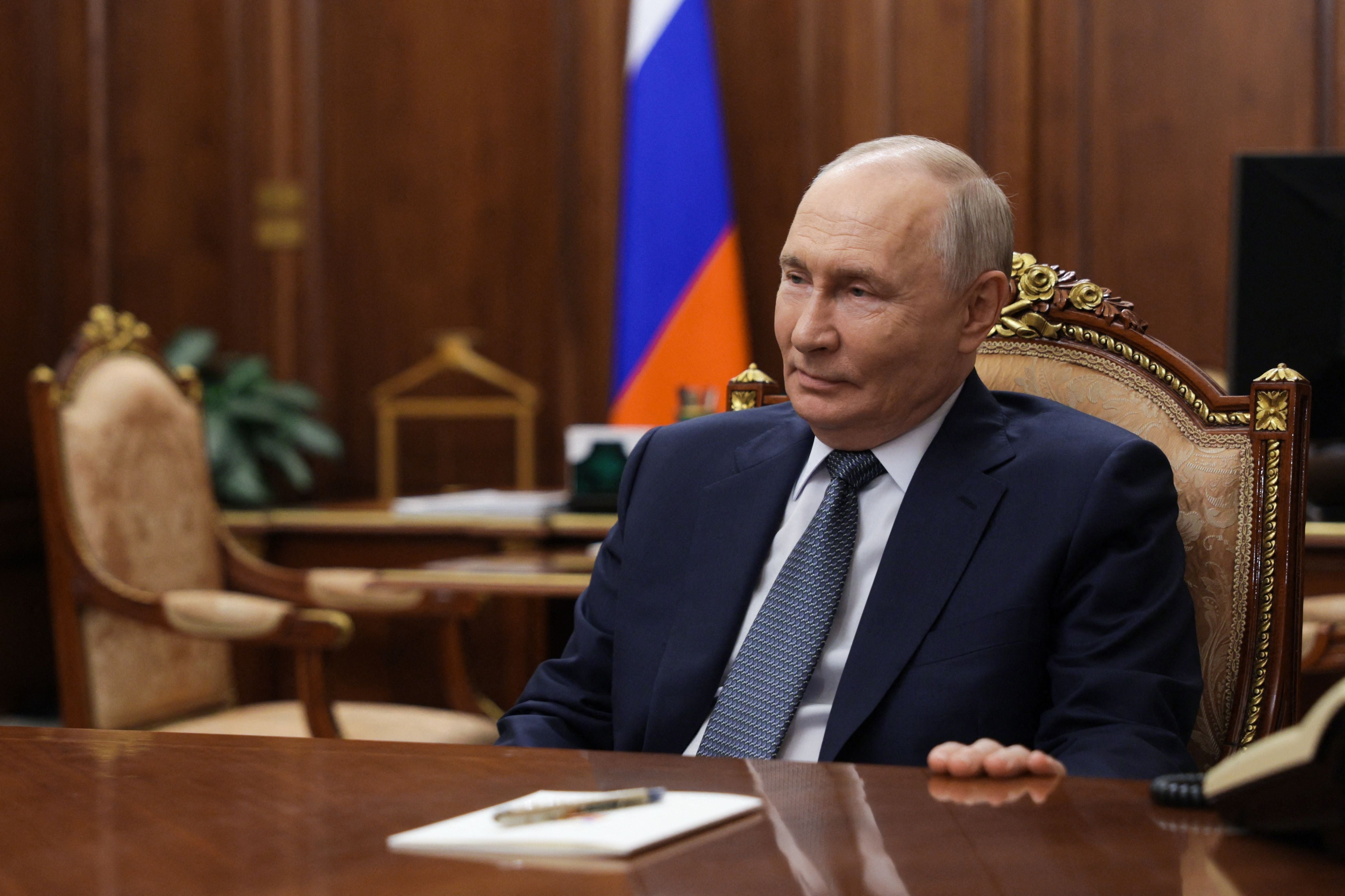 Ein Mann im Anzug sitzt an einem Tisch vor einer russischen Flagge in einem holzgetäfelten Raum.