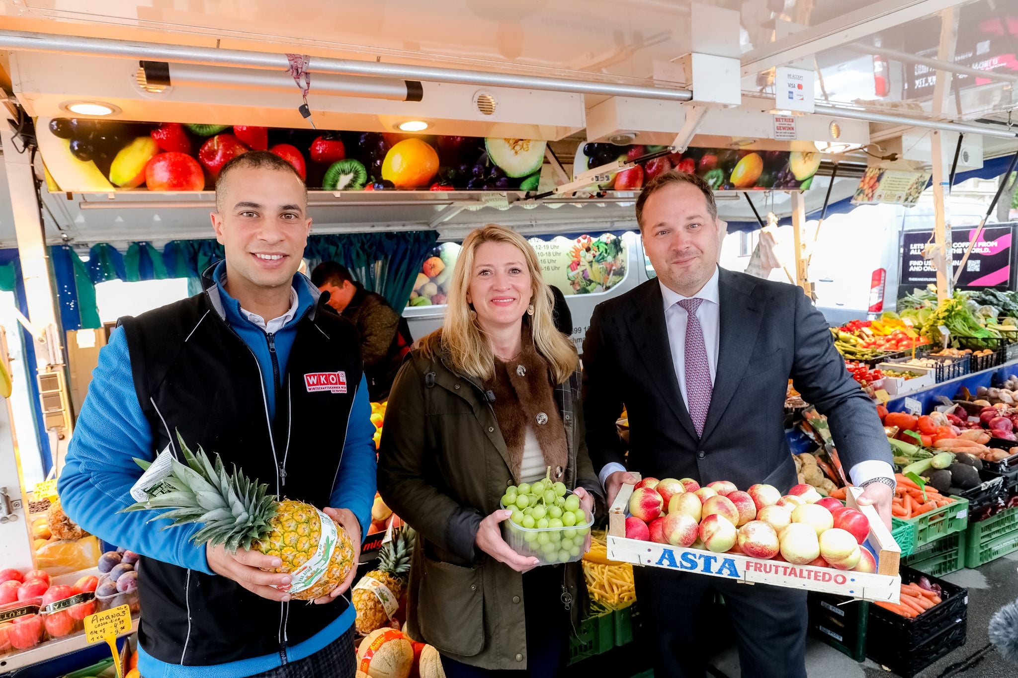 Drei Personen halten Ananas, Trauben und Äpfel auf einem Obst- und Gemüsemarkt.