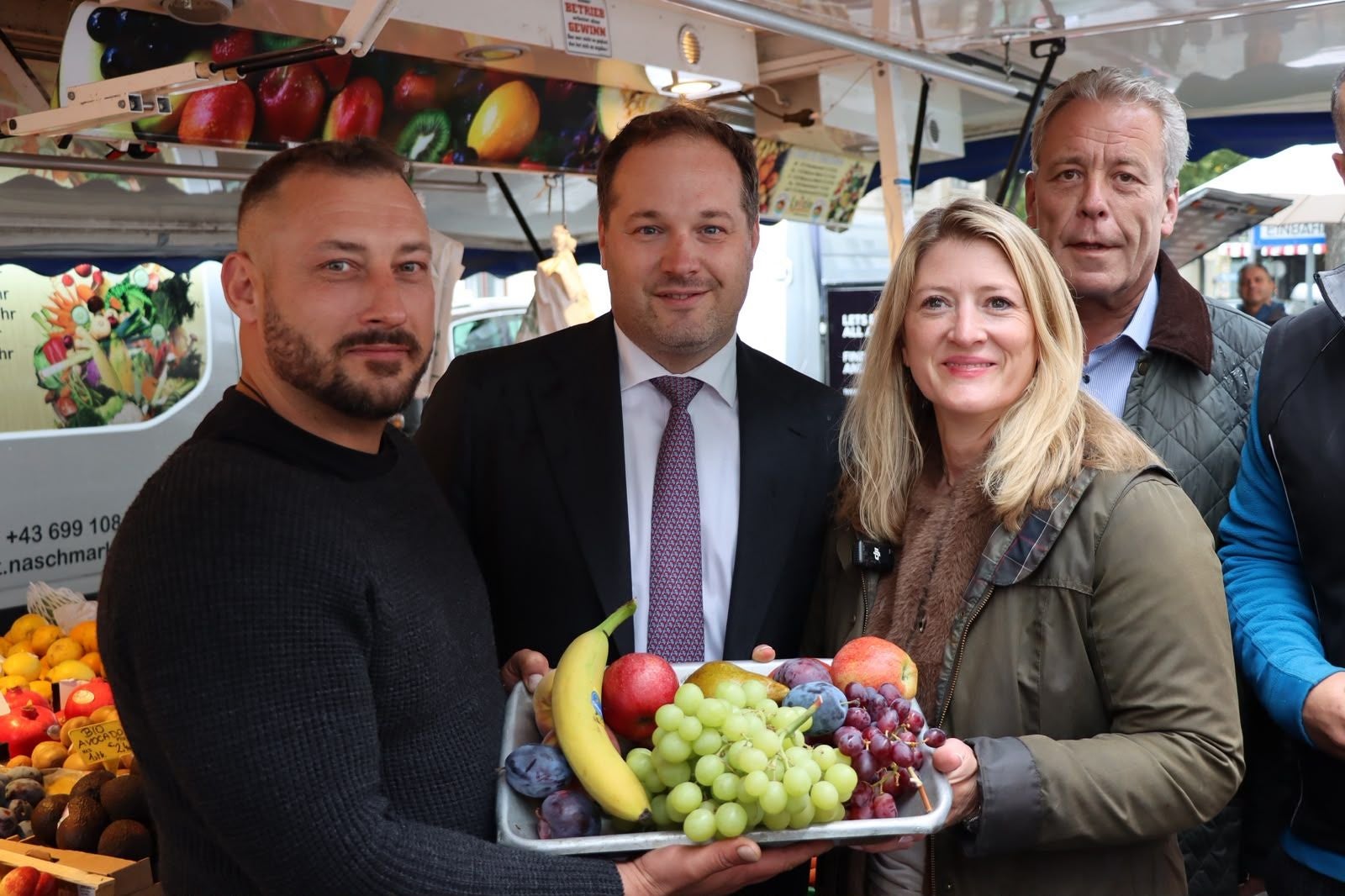 Drei Personen halten Ananas, Trauben und Äpfel auf einem Obst- und Gemüsemarkt.
