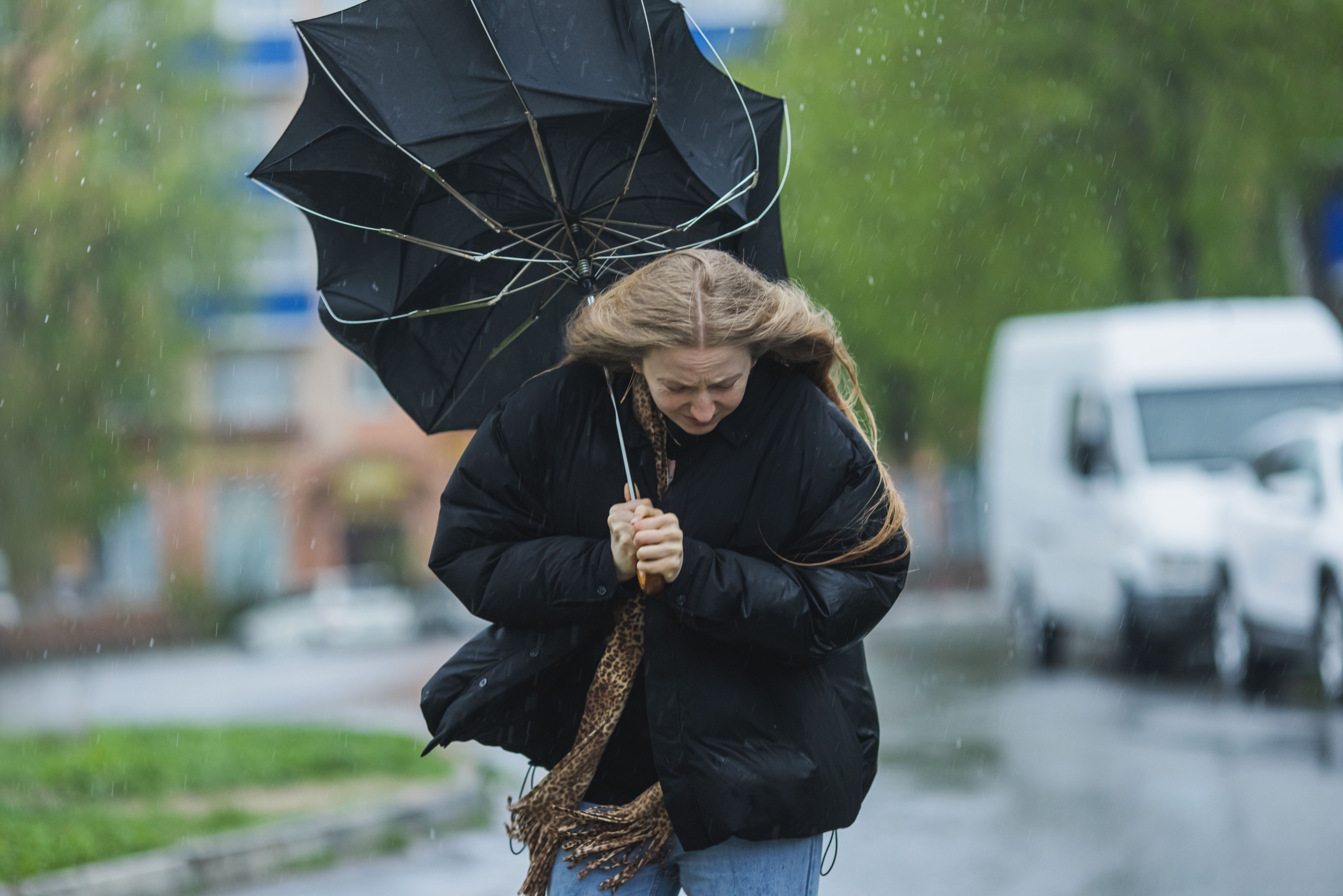 Frau kämpft bei Sturm und Regen mit umgeklapptem Regenschirm auf einer Straße.