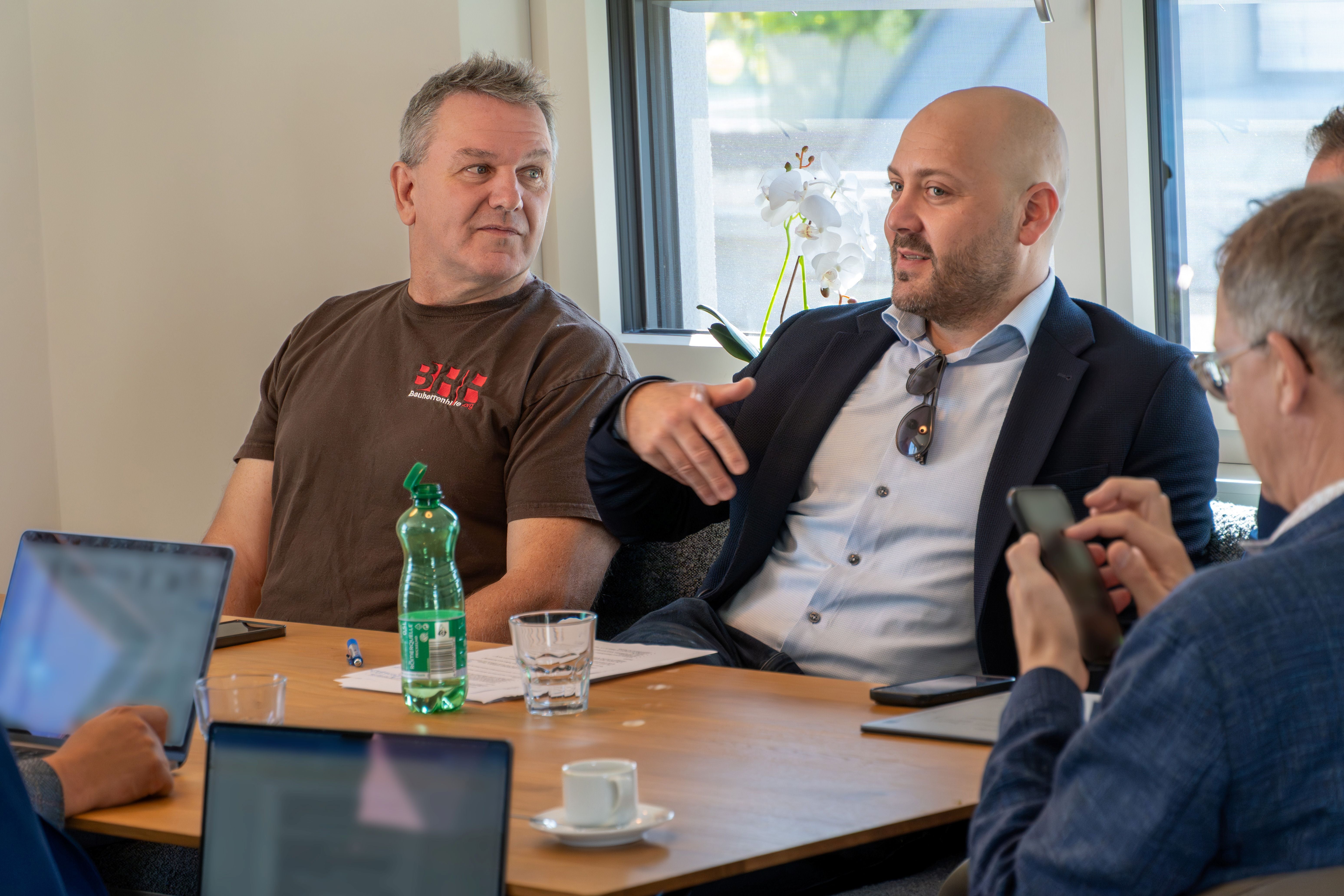 Vier Männer sitzen an einem Tisch und sprechen miteinander, Laptops und Getränke stehen davor.