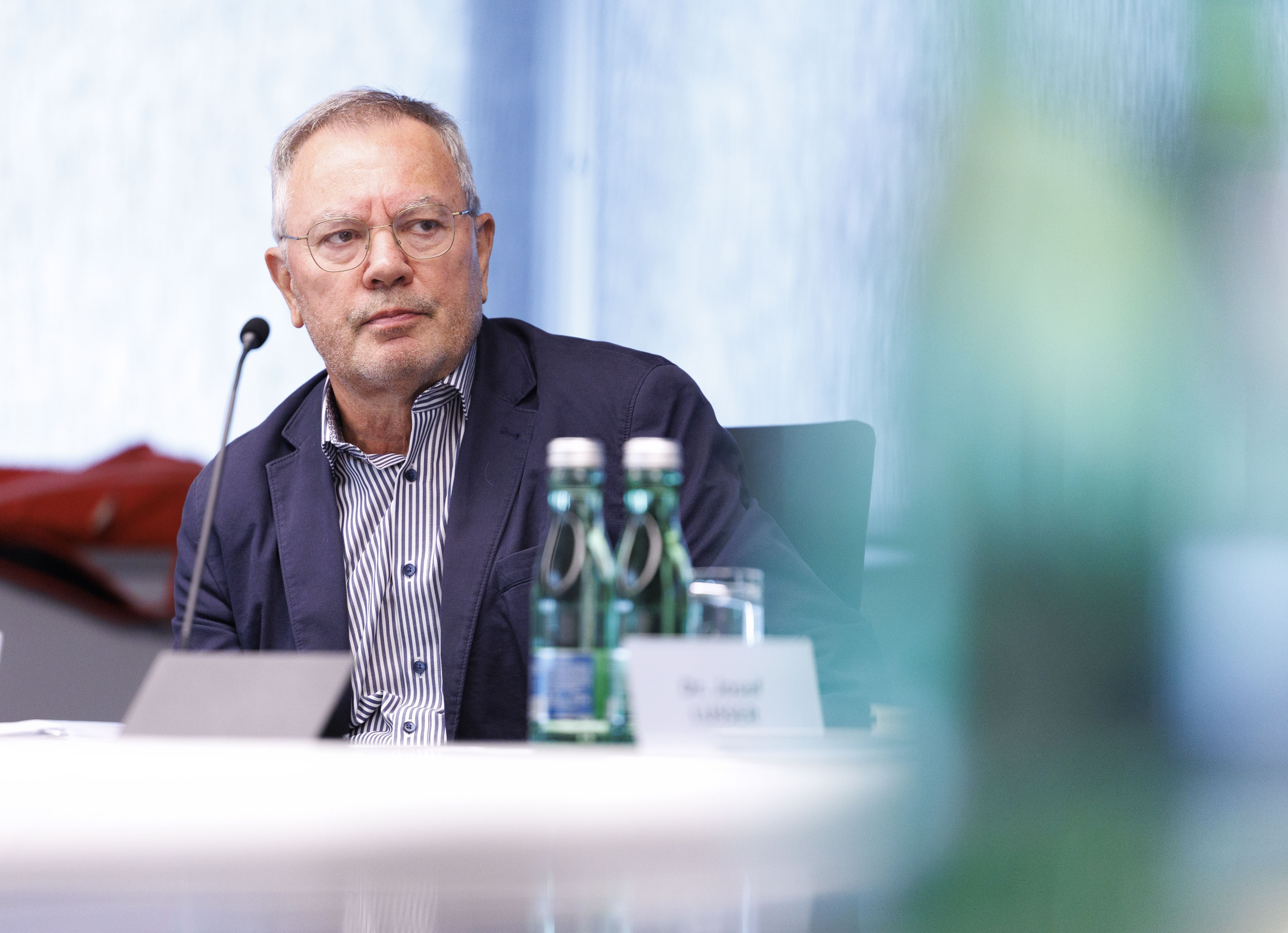Ein Mann mit Brille sitzt bei einer Konferenz am Tisch, vor ihm stehen Wasserflaschen.