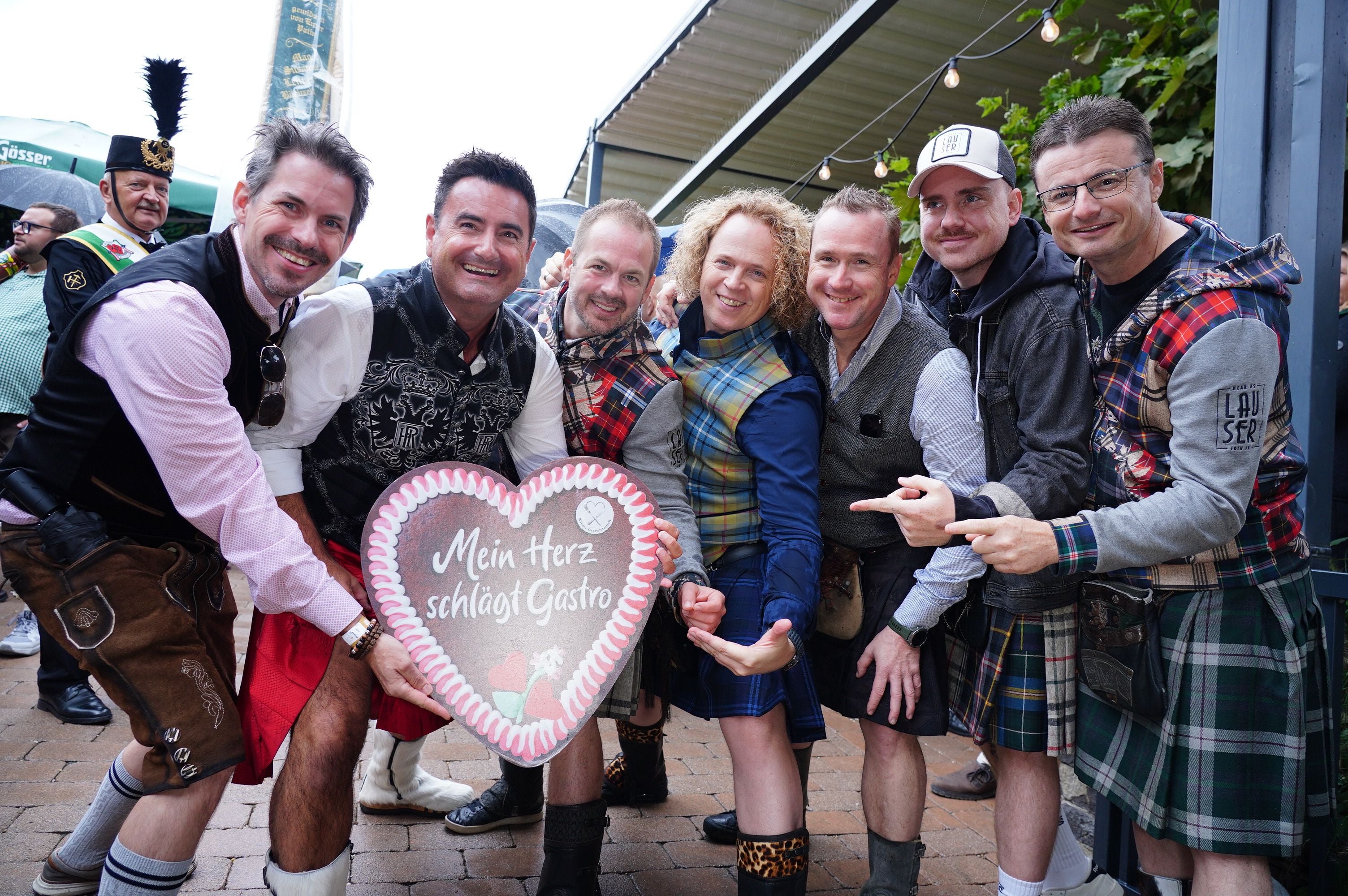Sieben Männer in Tracht posieren lachend mit einem Lebkuchenherz bei der Wiener Kaiser Wiesn.