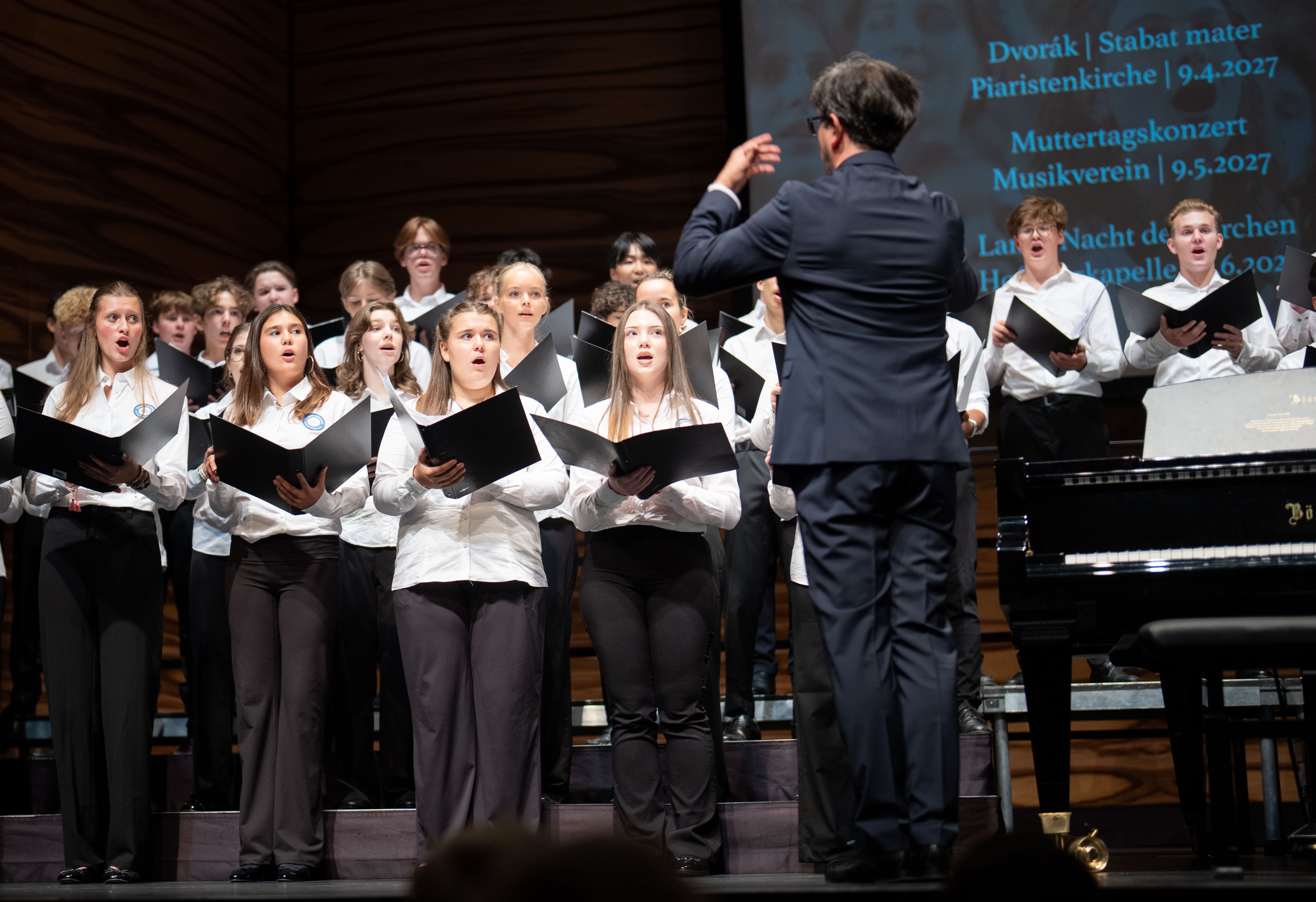 Ein Chor singt auf einer Bühne, dirigiert von einem Mann im Anzug, daneben ein Klavier.